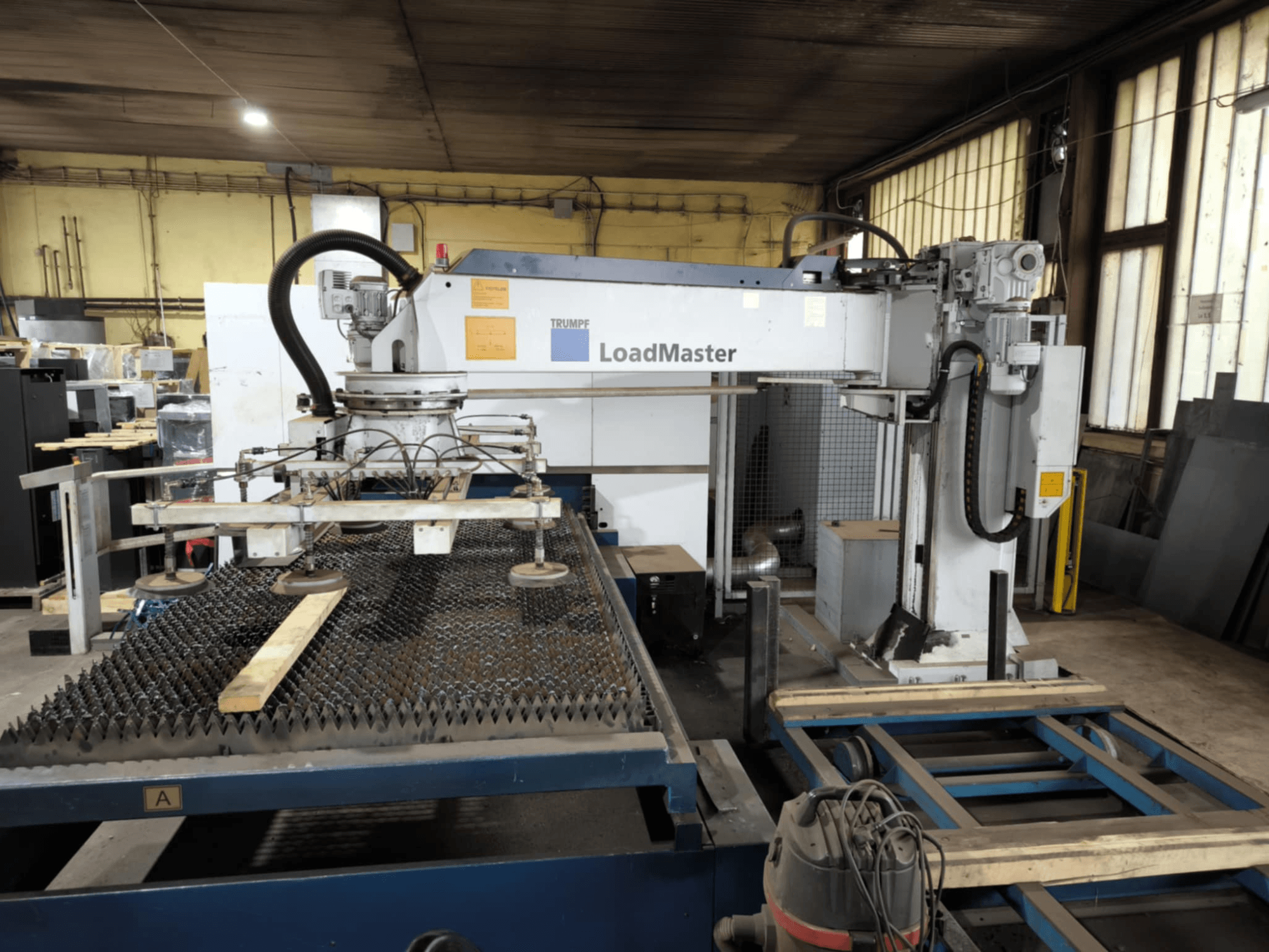TRUMPF TRULASER 3030 FIBER vágógép, elölnézetből, a LoadMaster rendszer bemutatásával, rácsos munkaasztalon elhelyezve.