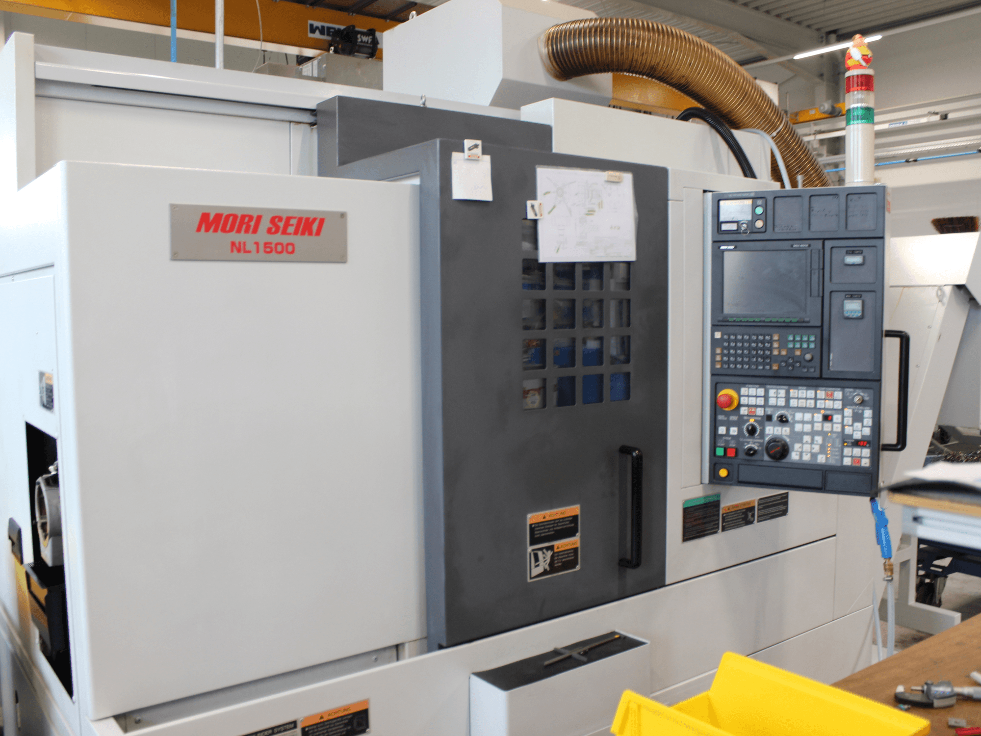 A MORI SEIKI NL 1500 SY eszterga-marógép oldalnézete, amelyen látható a vezérlőpanel, a munkavédelmi címkék és a hűtőrendszer.