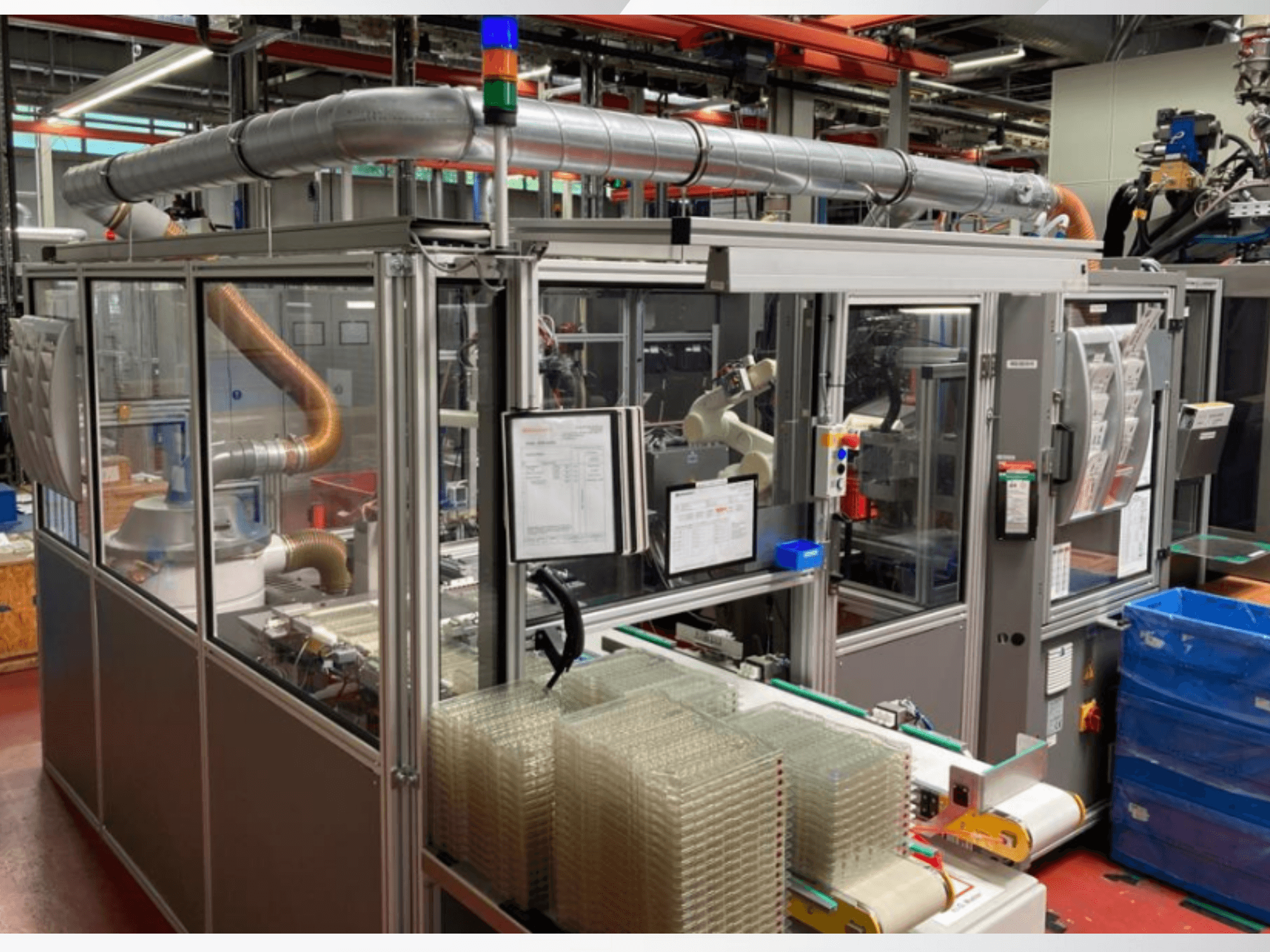Egy automatizált gép előlapja fémcsövekkel, vezérlőpanellel és egymásra helyezett átlátszó műanyag tartályokkal egy gyár padlóján.