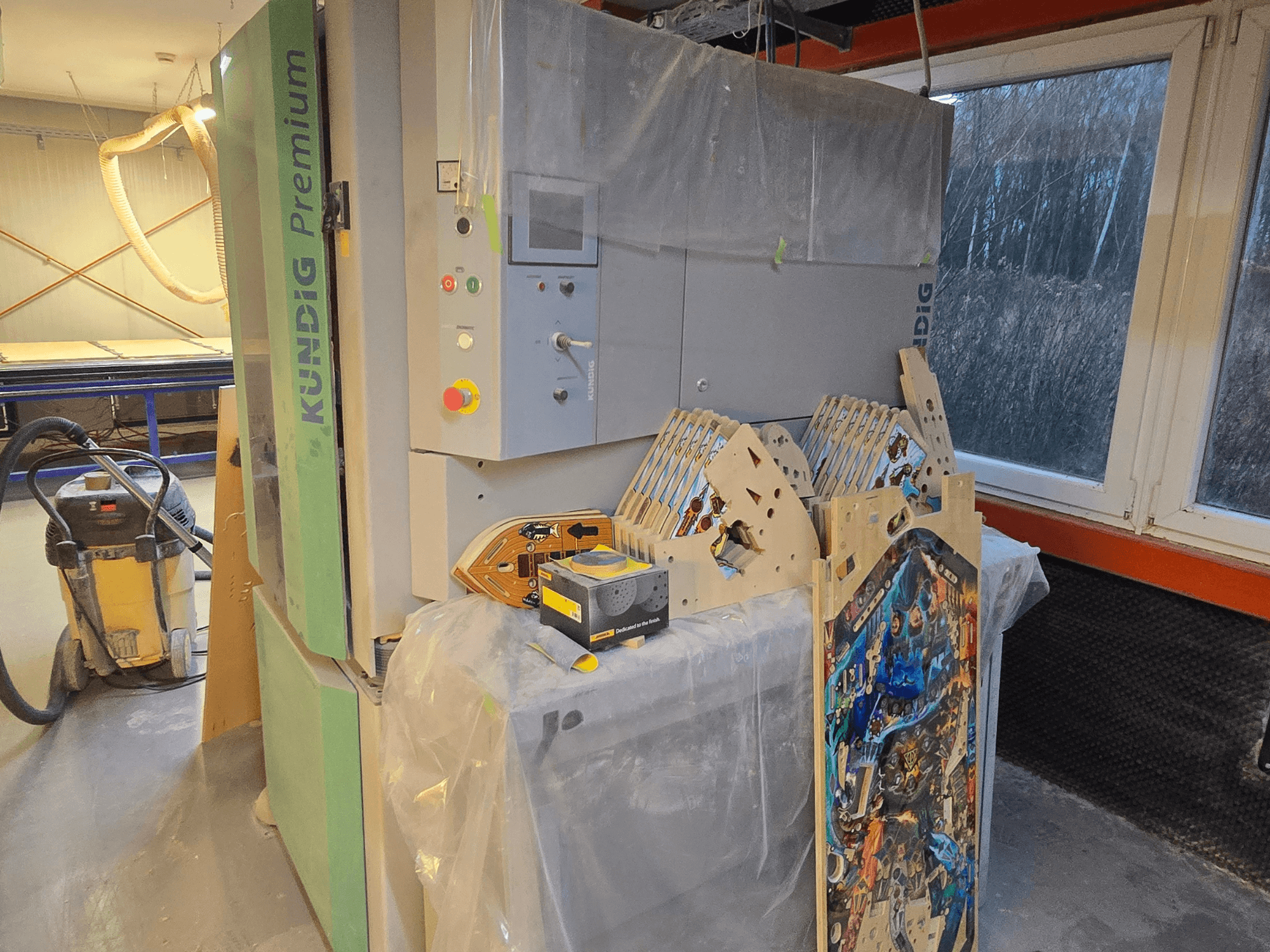 Kundig Premium-1 1100-C szalagcsiszoló műhelyben, a kezelőpanel és a fadarabok oldalnézetben, a háttérben porszívóval.