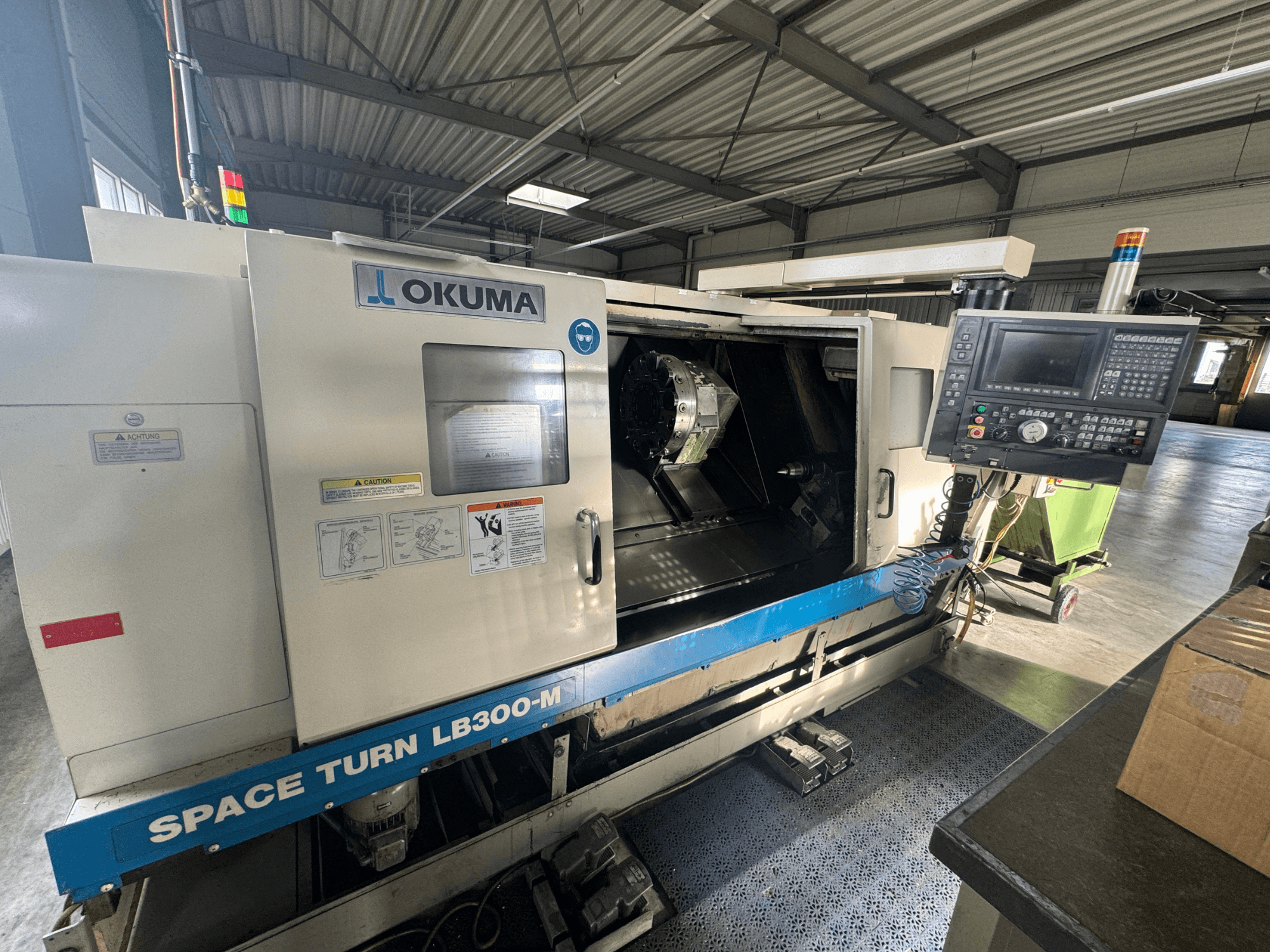 Okuma Space Turn LB300-M CNC eszterga elölnézetben, a vezérlőpanel és a nyitott ajtó, amely a belső megmunkálási területet mutatja.