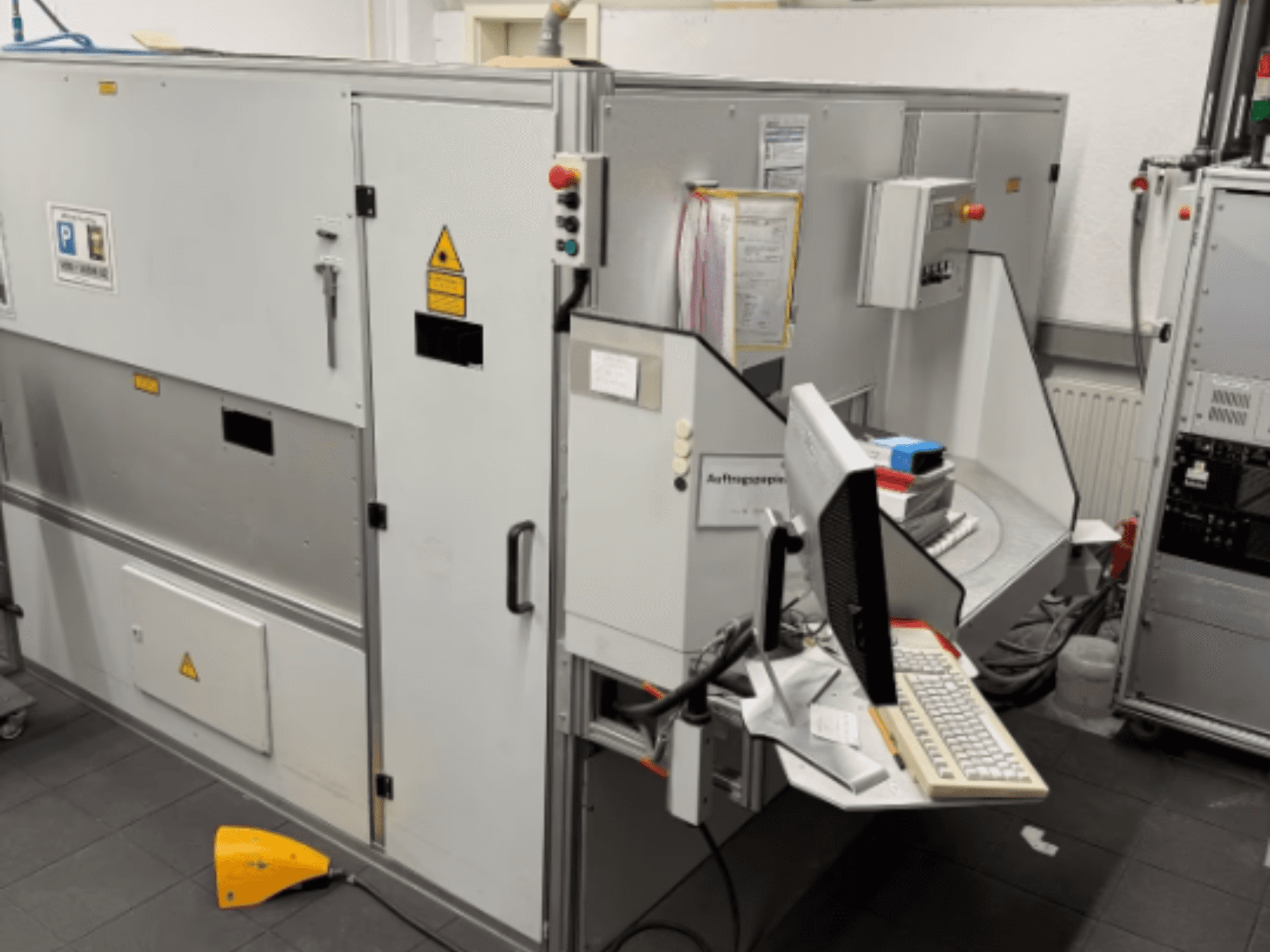 Ipari gép számítógépes terminállal, oldalnézet; vezérlőgombokkal és biztonsági figyelmeztető táblákkal.