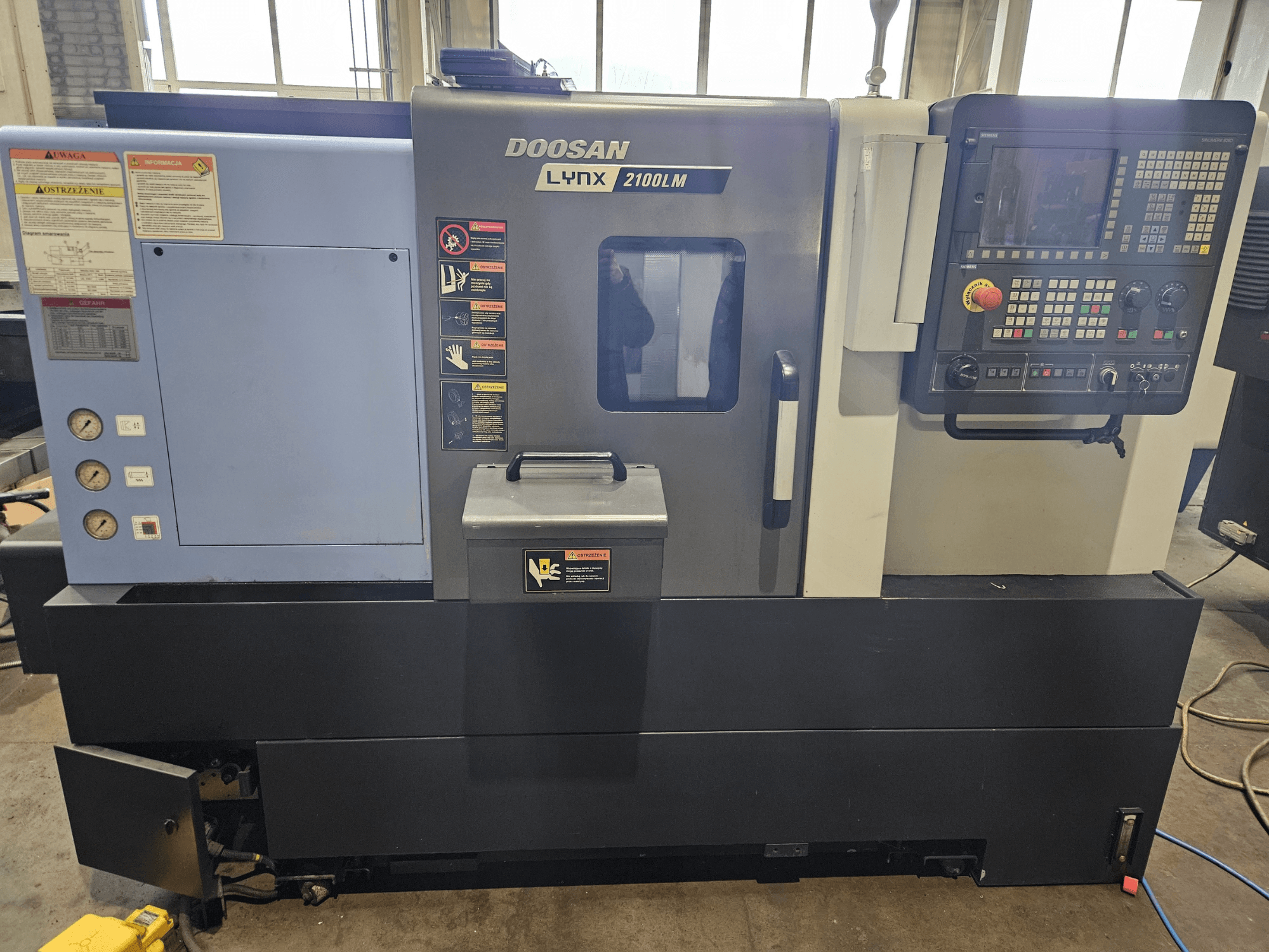 Doosan Lynx 2100LM CNC eszterga, elölnézet, a vezérlőpanel, a légnyomásmérők és a biztonsági címkék láthatóak.