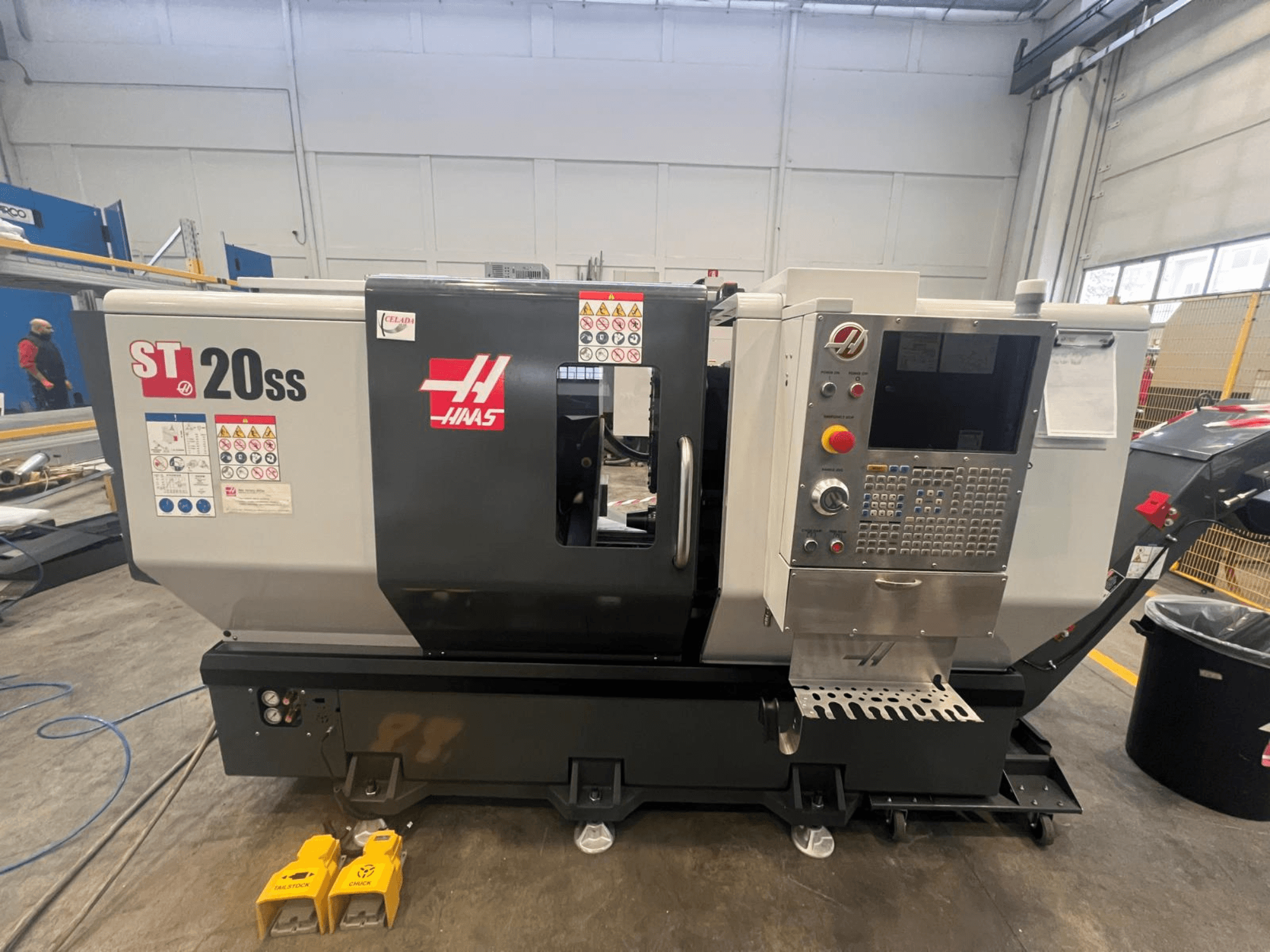 Haas ST 20SS CNC esztergapad oldalnézetben, a vezérlőpanel, a biztonsági címkék és a padlón lévő lábpedálok.