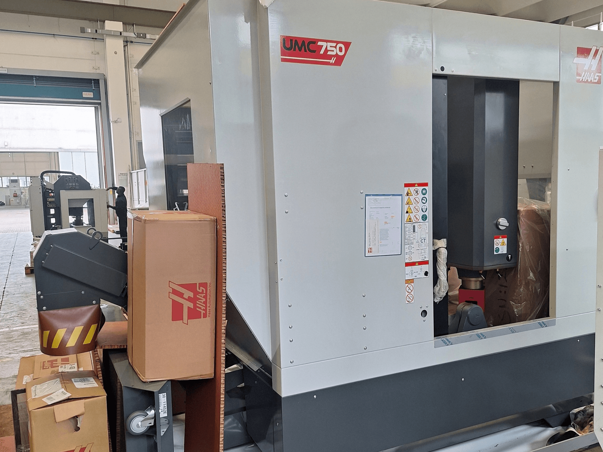 HAAS UMC-750 univerzális megmunkáló központ, oldalnézet, amelyen látható a vezérlőpanel és a szerszámtároló figyelmeztető címkékkel és csomagolással együtt.