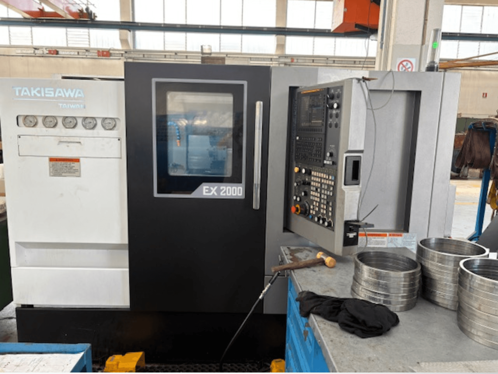 Takisawa EX 2000 CNC-gép, elölnézet, vezérlőpanel, műszerek és fém alkatrészek egy műhelyasztalon.