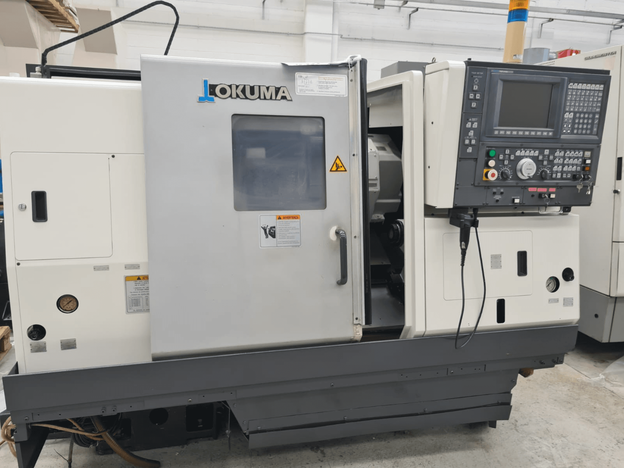Okuma LB 300M X 500 vízszintes esztergagép oldalnézetben, a vezérlőpanel, az orsóhoz való hozzáférés és a biztonsági figyelmeztetések bemutatásával.