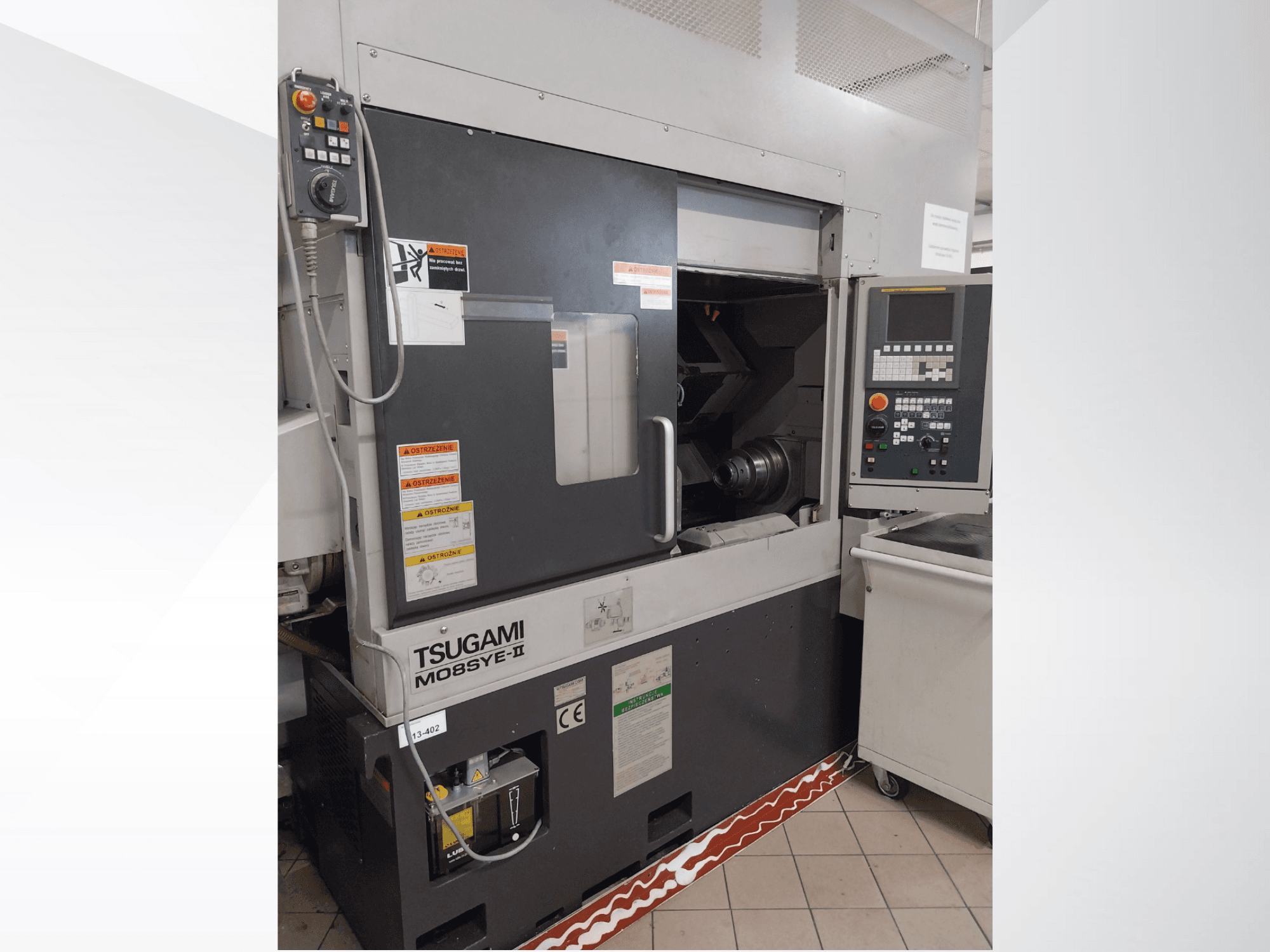 Tsugami M08SY-II CNC eszterga, előlnézet, a vezérlőpanel, az orsó és a betöltőterület látható. Biztonsági figyelmeztetések és márkajelzés.