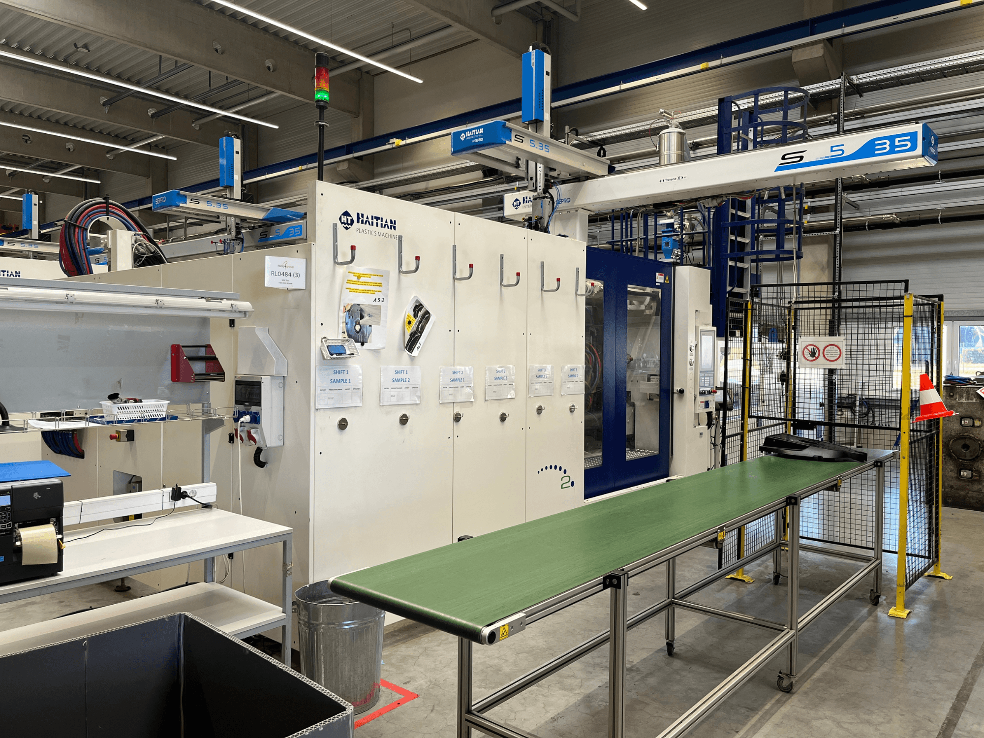 Haiti S5-35 műanyagipari gép egy műhelyben; zöld szállítóasztal, biztonsági korlátok és a vezérlőpanel látható elölnézetből.