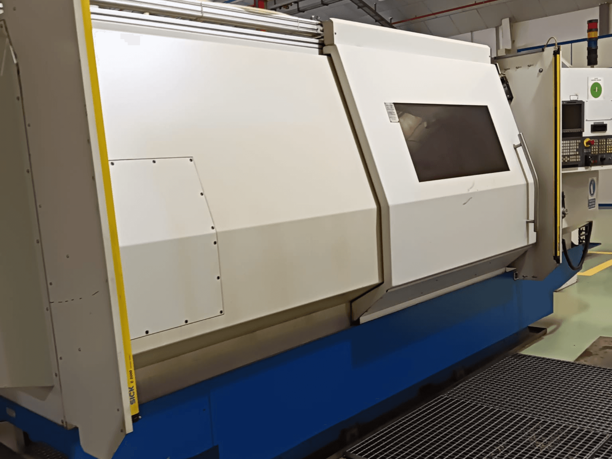 Ipari esztergagép oldalnézetben, nagy fehér és kék burkolattal, digitális vezérlőpanellel és biztonsági berendezésekkel.