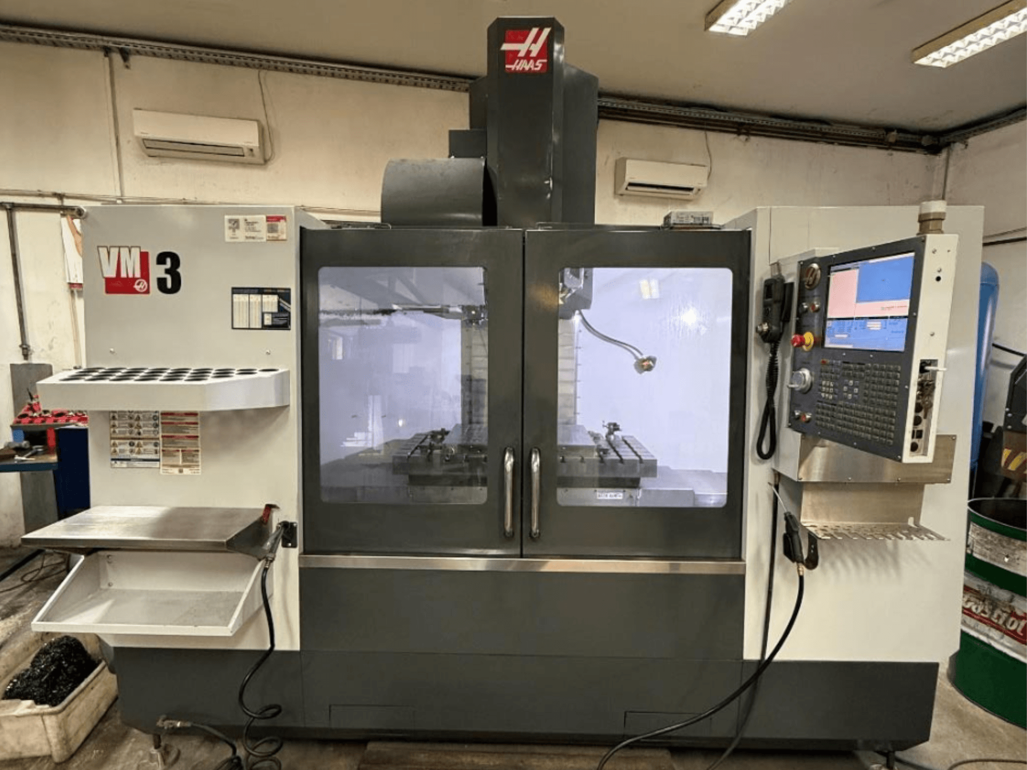 A Haas VM-3 függőleges megmunkáló központ elölnézetben, nagy vezérlőpanellel és a szerszámokkal ellátott, jól látható munkaterülettel.