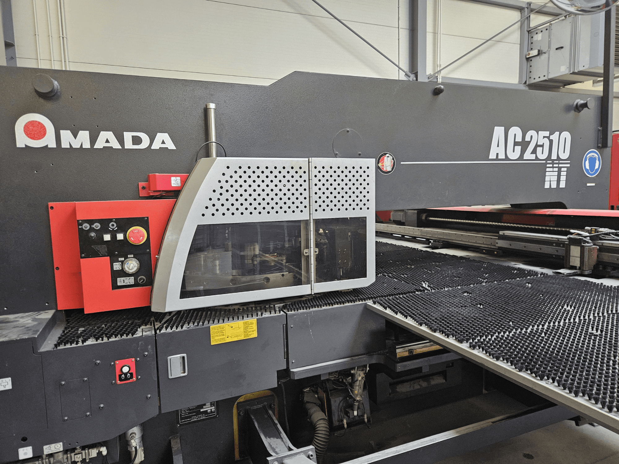 Amada AC2510 NT lézervágógép, előlnézet, a kezelőpanel gombokkal, gombokkal és biztonsági fedéllel.