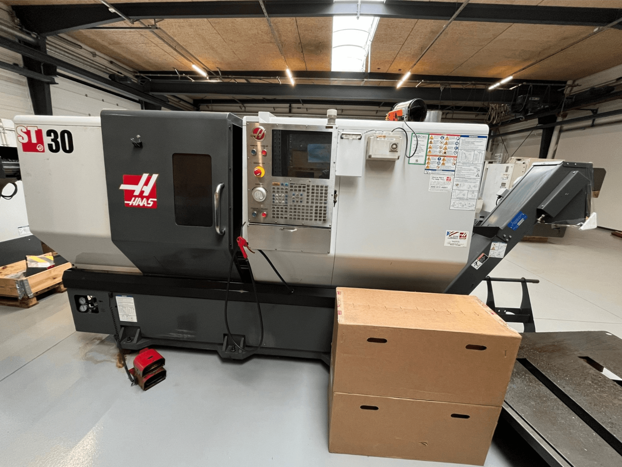 Haas ST-30 CNC eszterga, elölnézet, vezérlőpanel gombokkal, vészleállítóval és két egymásra helyezett kartondobozzal.