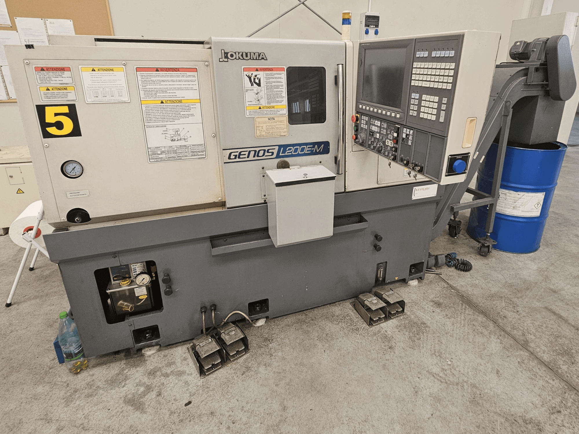 Okuma GENOS L200E-M vízszintes esztergagép elölnézetben, vezérlőpanel, címkék és hűtőfolyadékos hordó.