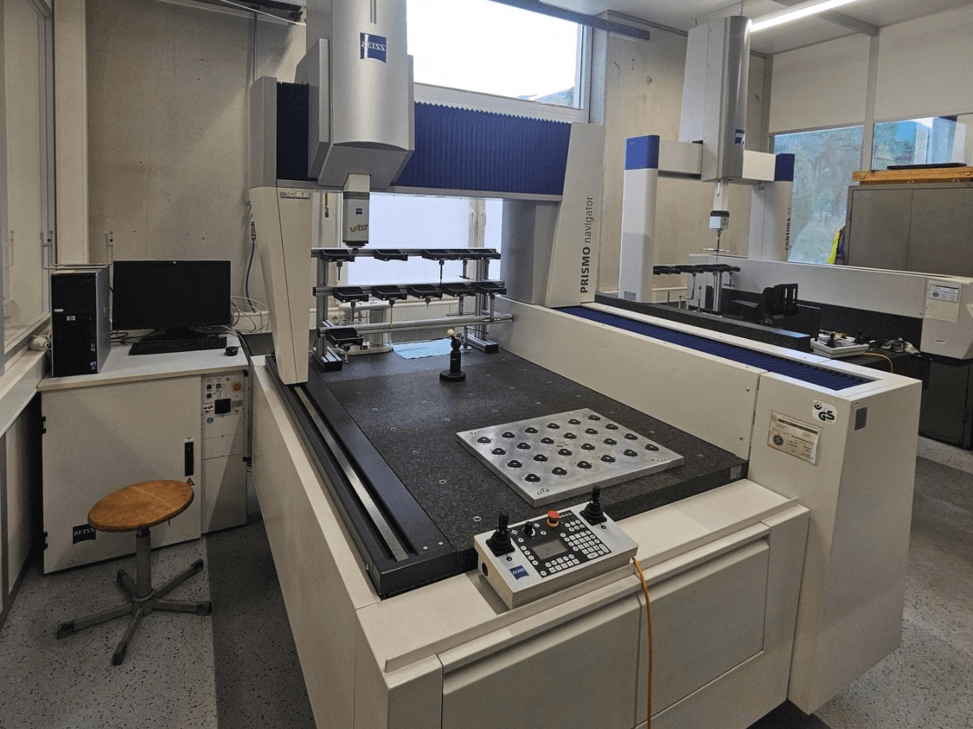 ZEISS Prismo Navigator 7 CMM egy laboratóriumban, elölnézetből, amelyen látható a vezérlőpanel, a munkaasztal és a monitor.