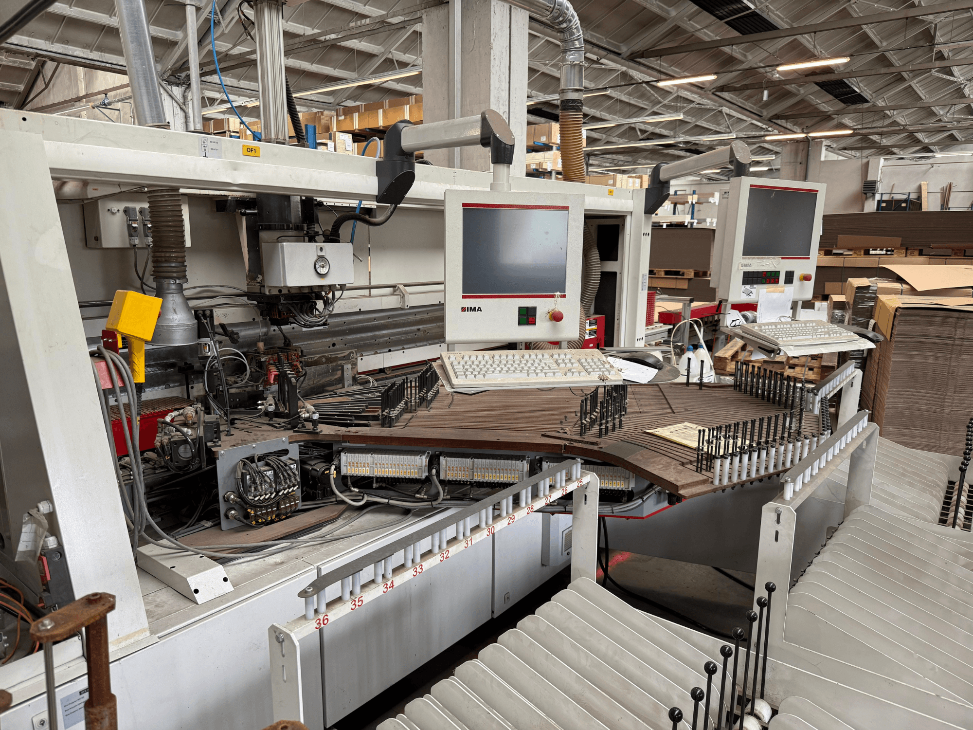 IMA precíziós famegmunkáló gép kettős monitorral és vezérlőpanellel, elölnézetben a szállítószalag és a szerszámterület.
