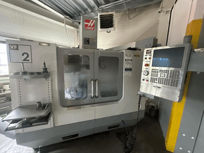 Haas VF-2 CNC-gép, elölnézet, a vezérlőszervekkel, a szerszámtartóval és az átlátszó ajtókkal, amelyeken a szerszámok a szekrény belsejében láthatók.
