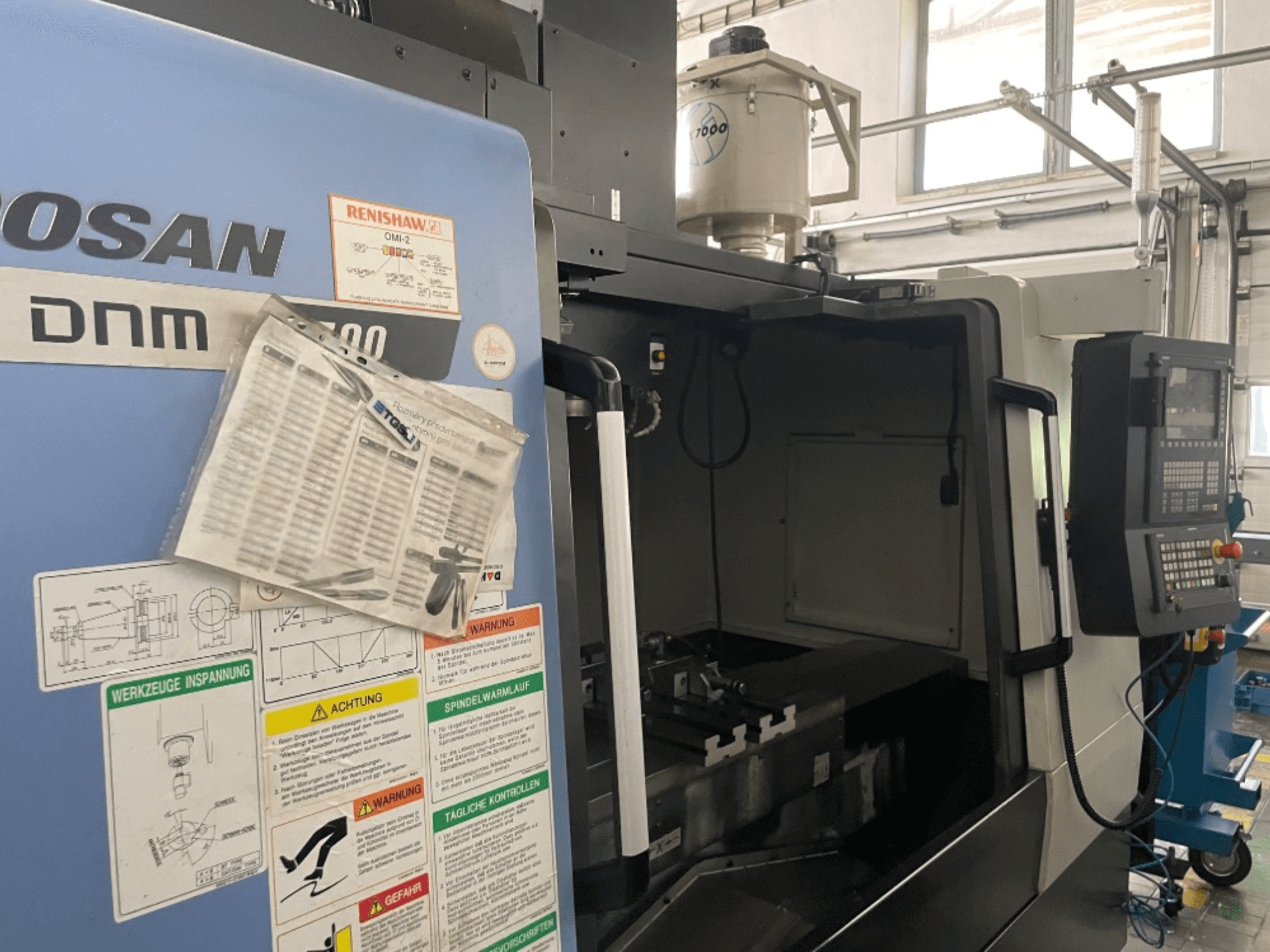 DOOSAN DNM 5700 függőleges megmunkálóközpont, előlnézet, a vezérlőpanel, a biztonsági címkék és a gép részletei.