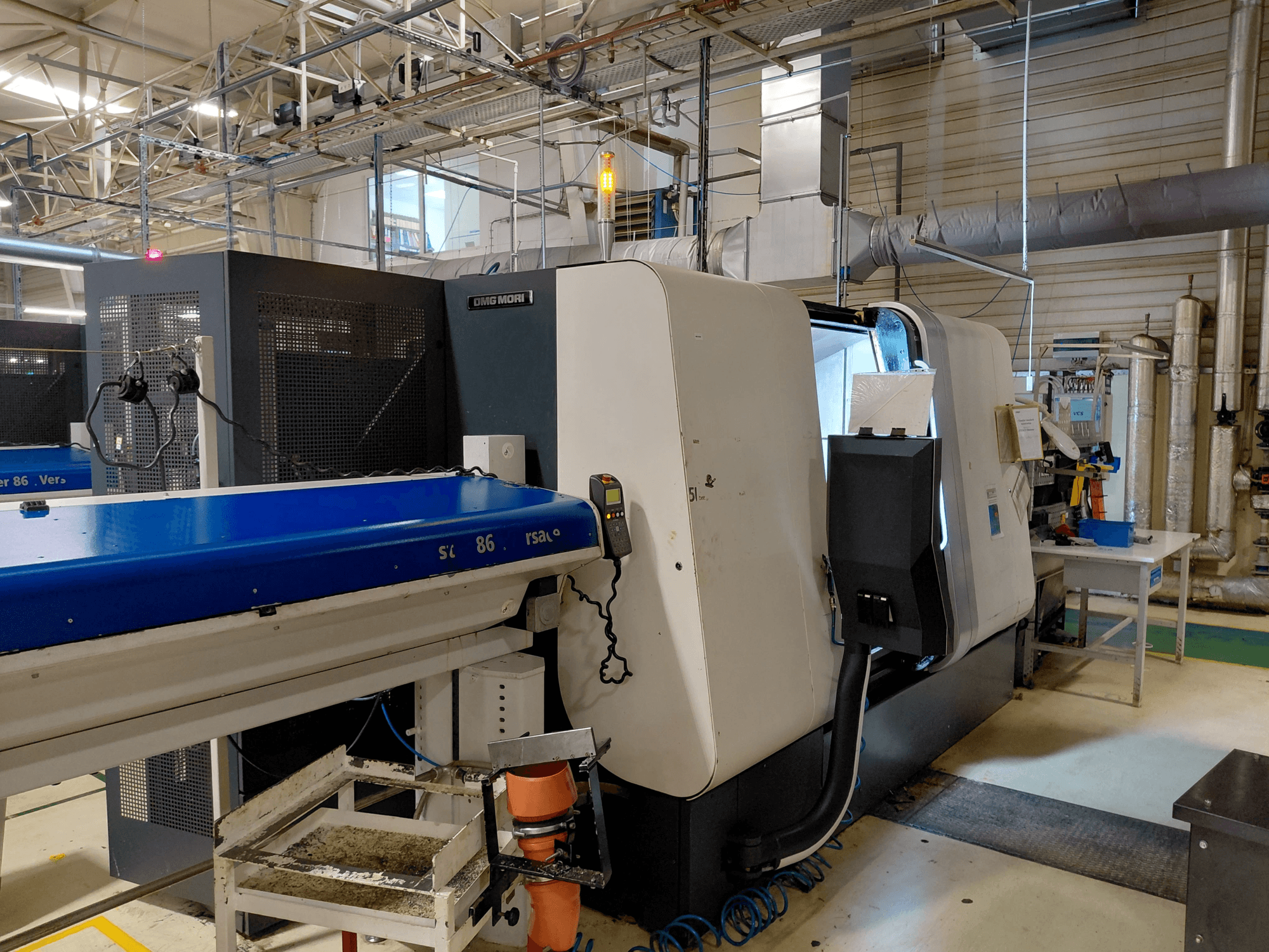 DMG MORI Sprint 65 lineáris többorsós esztergagép, oldalnézet a kezelőpanellel és a szállítószalaggal műhelyben.