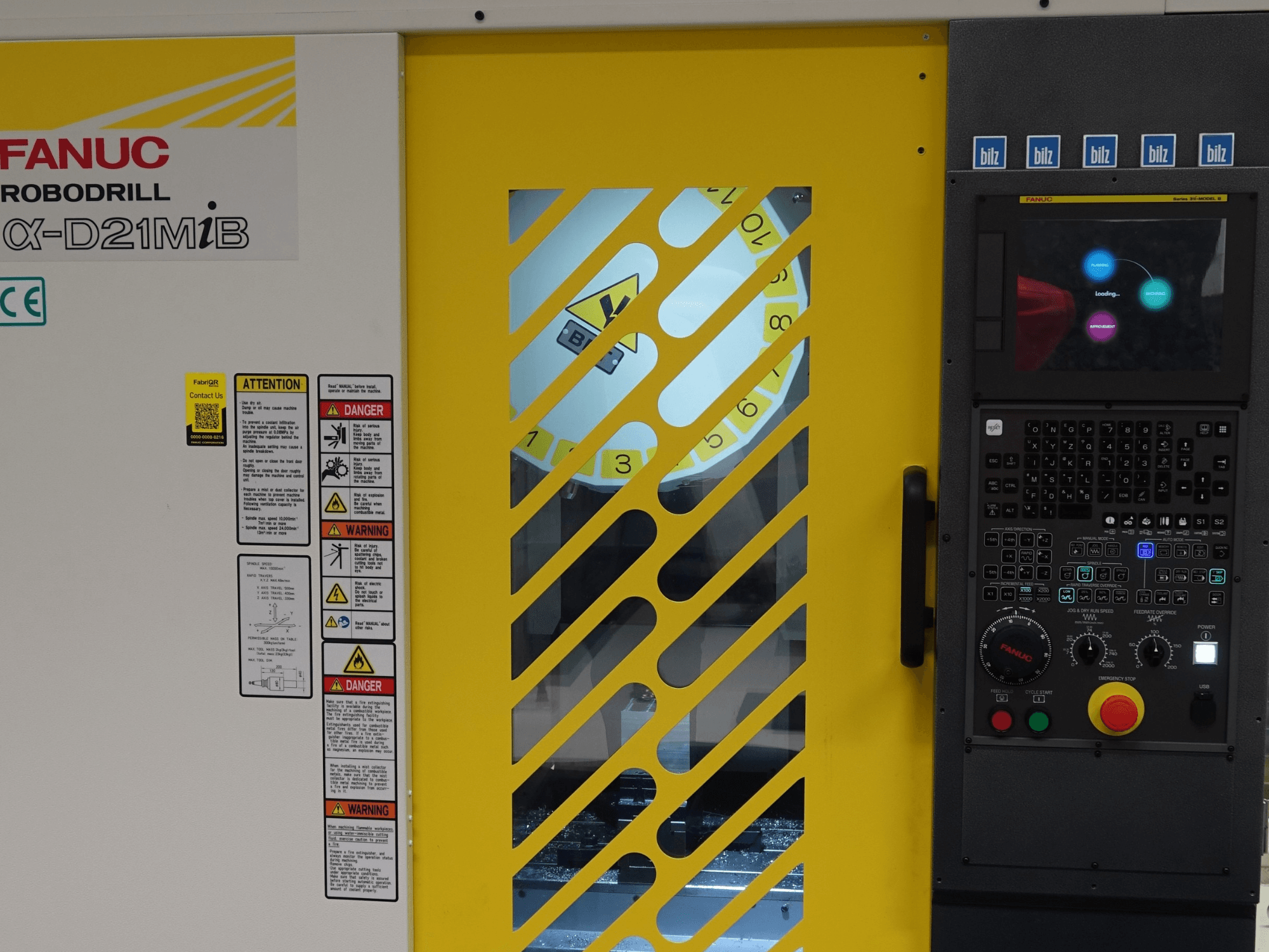 A FANUC α-D21MiB függőleges megmunkáló központ elölnézete, amelyen láthatók a biztonsági figyelmeztetések, a vezérlőpanel és a sárga biztonsági ajtó.