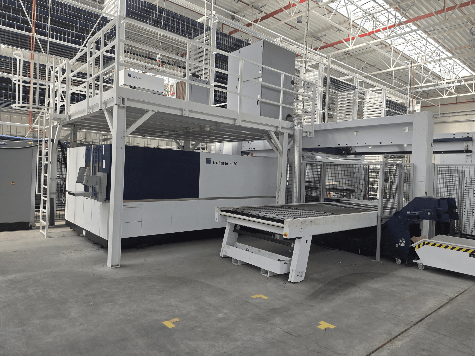 TRUMPF TruLaser 5030 fiber L76 FMC gép egy műhelyben, elölnézetből, amelyen látható a vágási terület és a be- és kirakó mechanizmus.