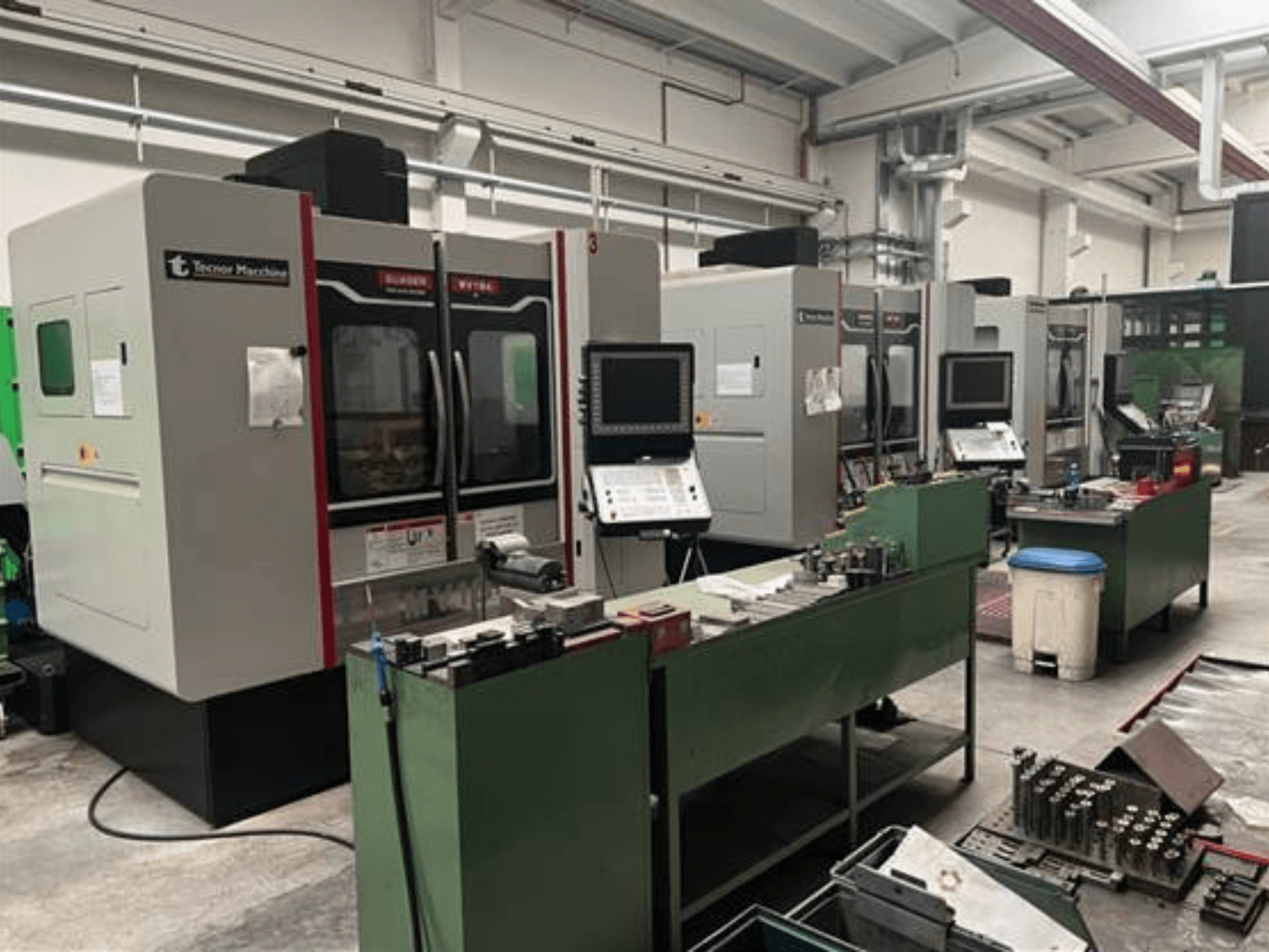 Négy CNC-gép a Tecnoer Marchine-tól egy műhelyben, elölnézetben, szerszámokkal és alkatrészekkel a zöld munkapadokon.