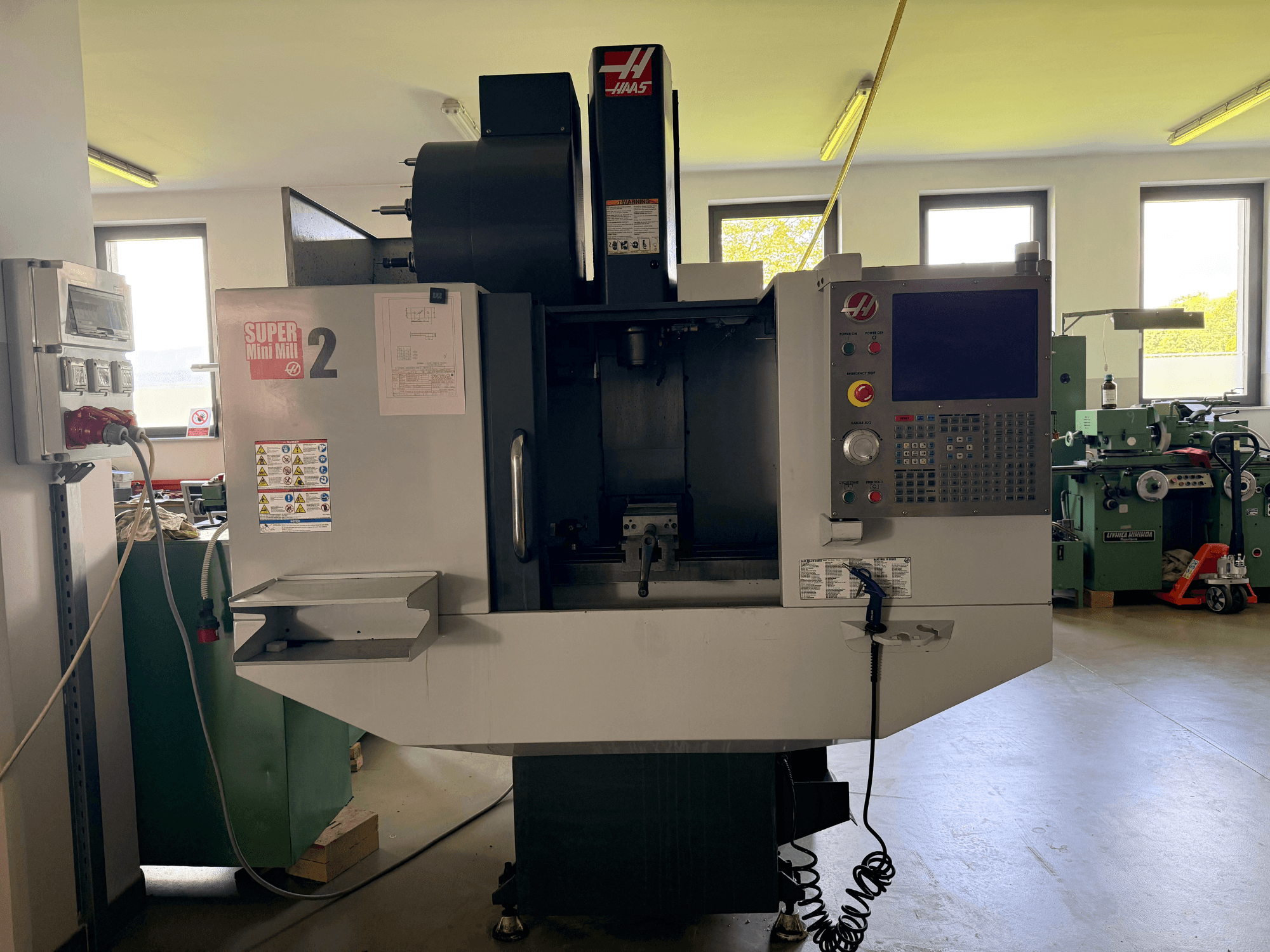 Haas Mini Mill 2 függőleges megmunkálóközpont elölnézetben, a vezérlőpanel és a szerszámterület figyelmeztető feliratokkal.