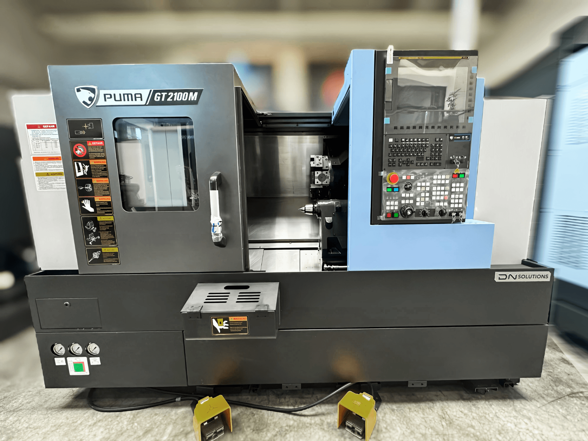 DN SOLUTIONS PUMA GT2100M esztergapad-központ, elölnézetben a vezérlőpanel, a megmunkáló kamra és a biztonsági címkék.