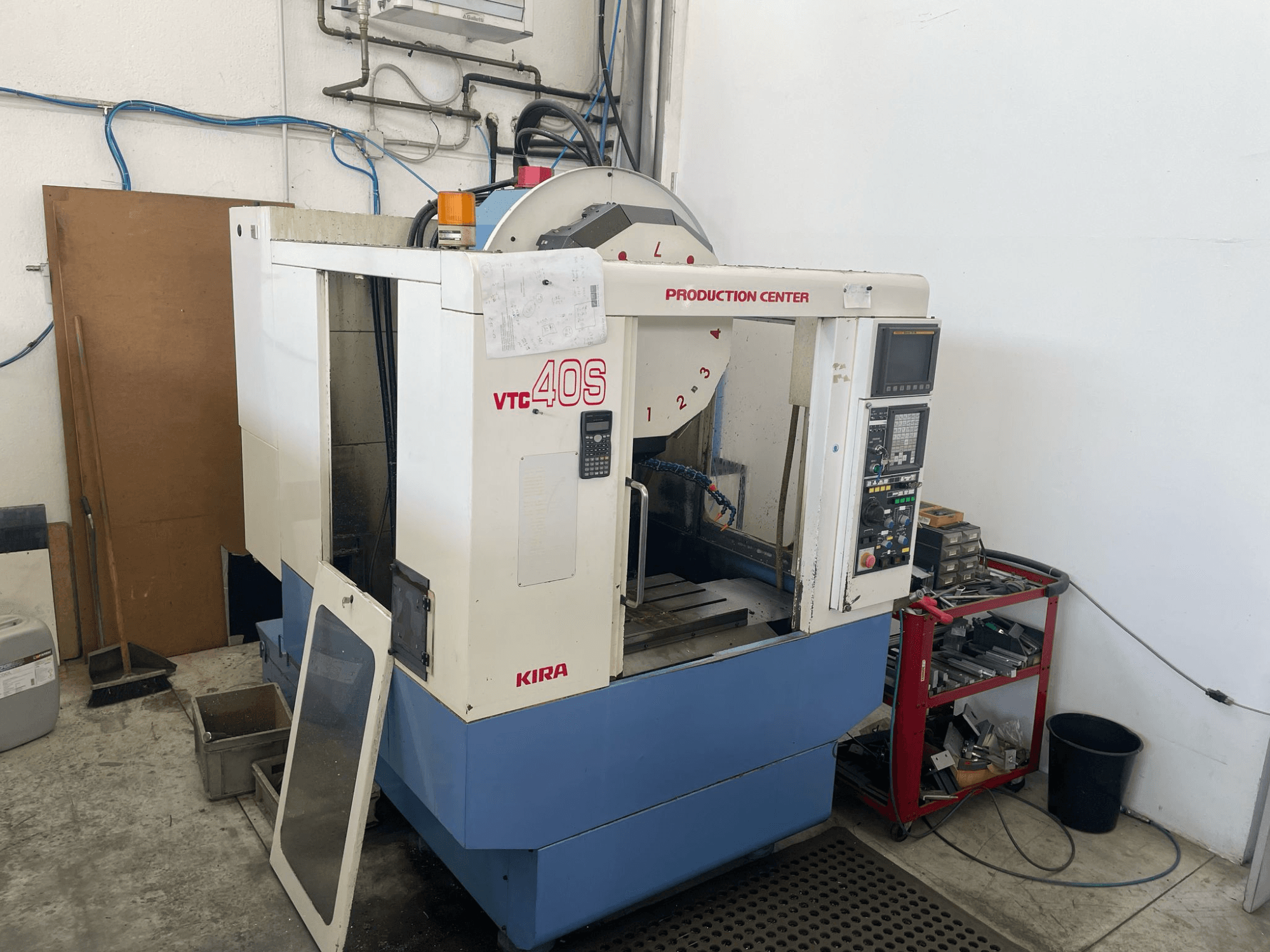 KIRA VTC 40S megmunkálóközpont oldalnézetben, vezérlőpanel, nyitott ajtó és szerszámok a közeli tárolókocsin.