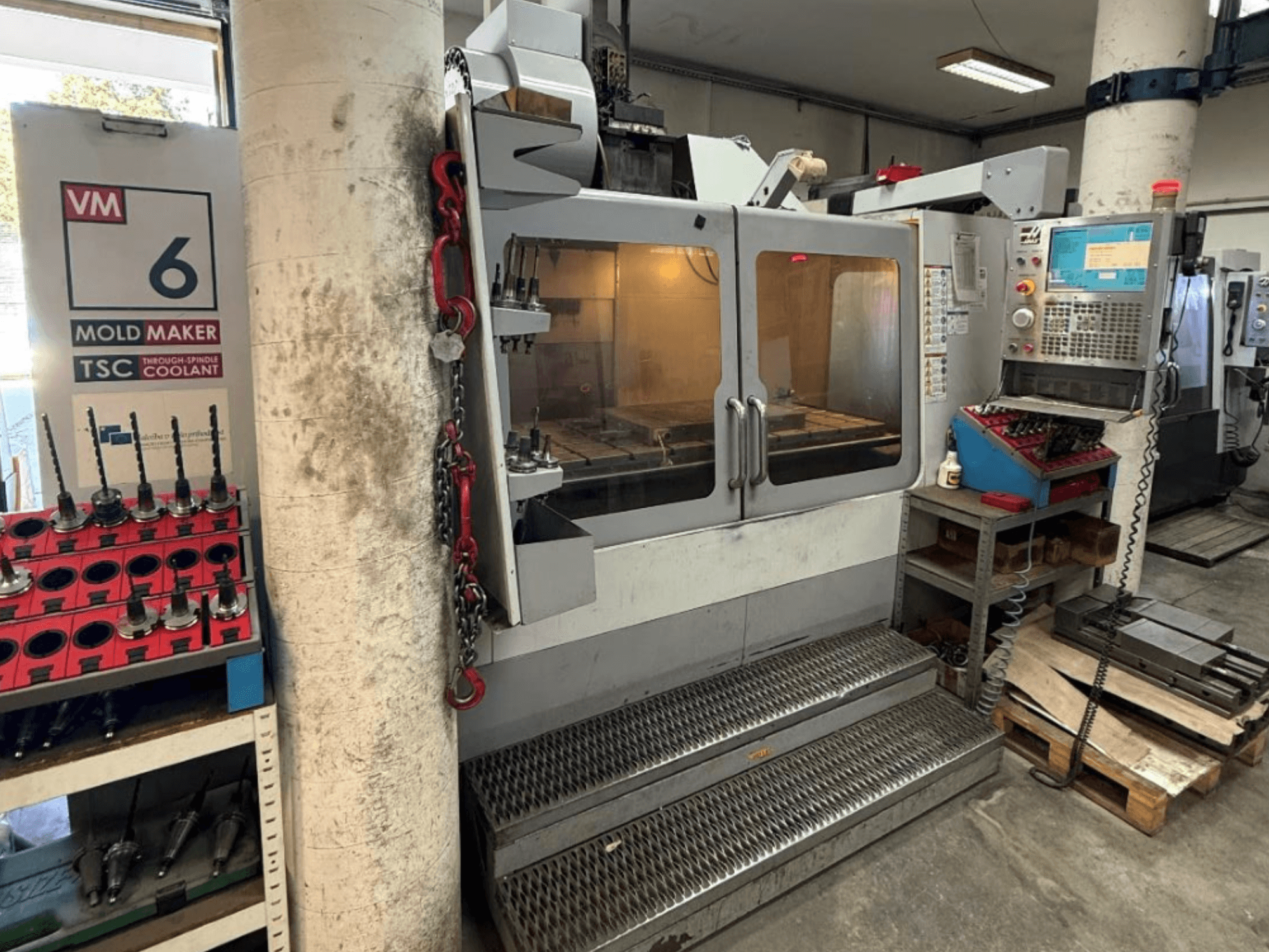 A HAAS VM-6 HE függőleges megmunkáló központ elölnézete, amelyen láthatók a kezelőszervek, a hűtőfolyadék-rendszer és a szerszámtároló.