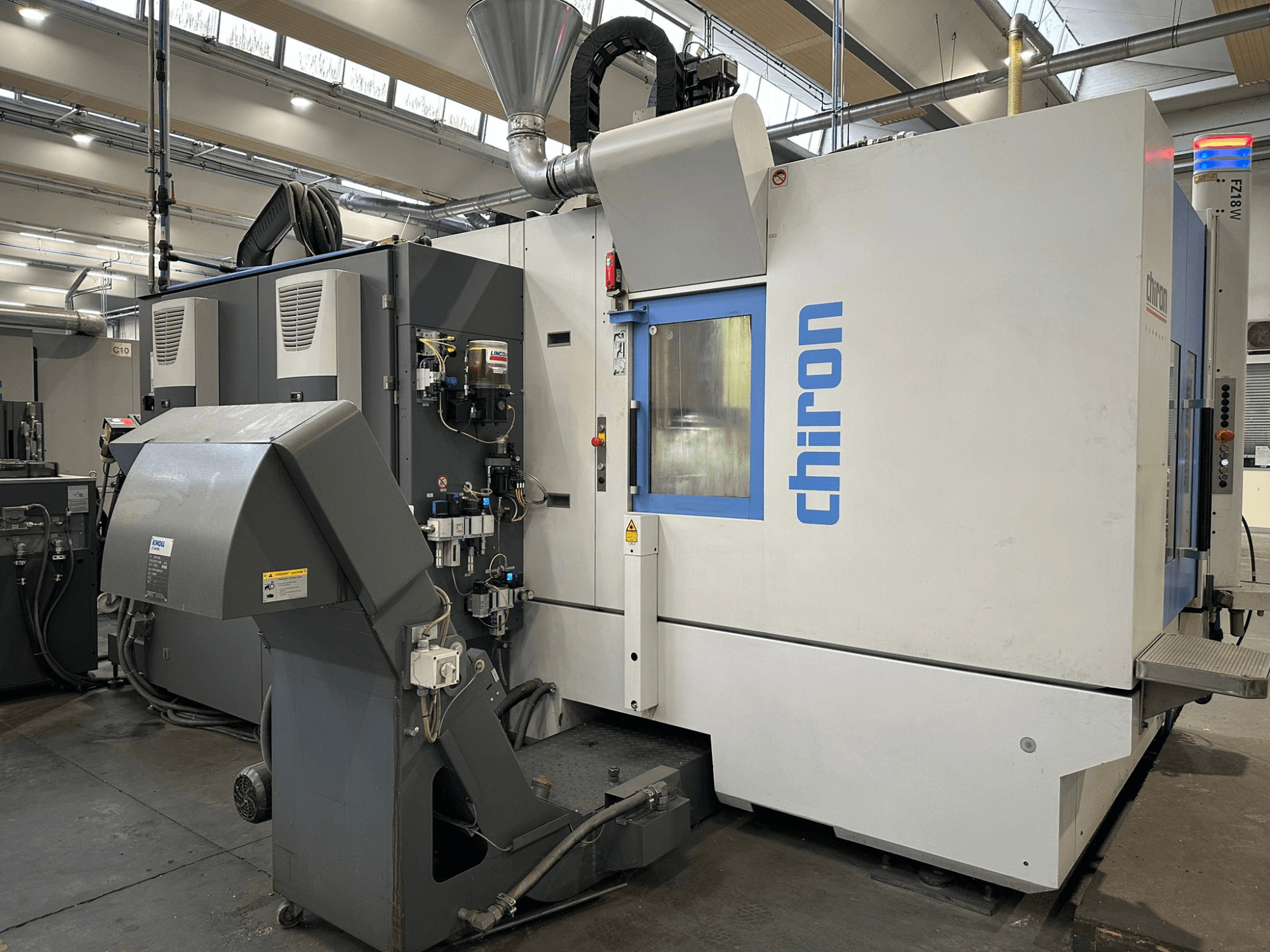 Chiron CNC-gép szürke és fehér színben, elölnézetben, kezelőszervekkel, csövekkel és porelszívó motorháztetővel.