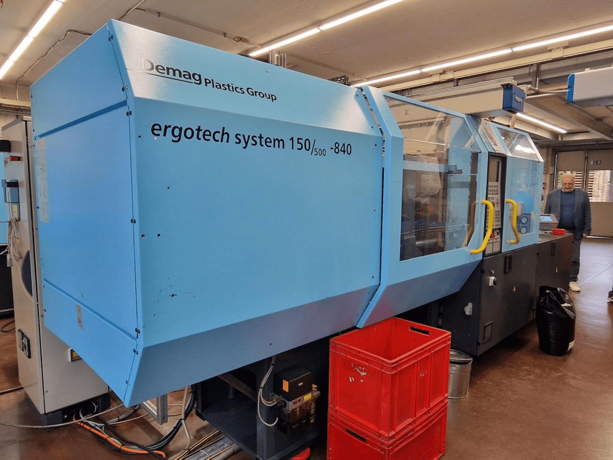 Demag Ergotech System 150/500-840 fröccsöntőgép kék színben, oldalnézetben, ládákkal és kezelővel a háttérben.