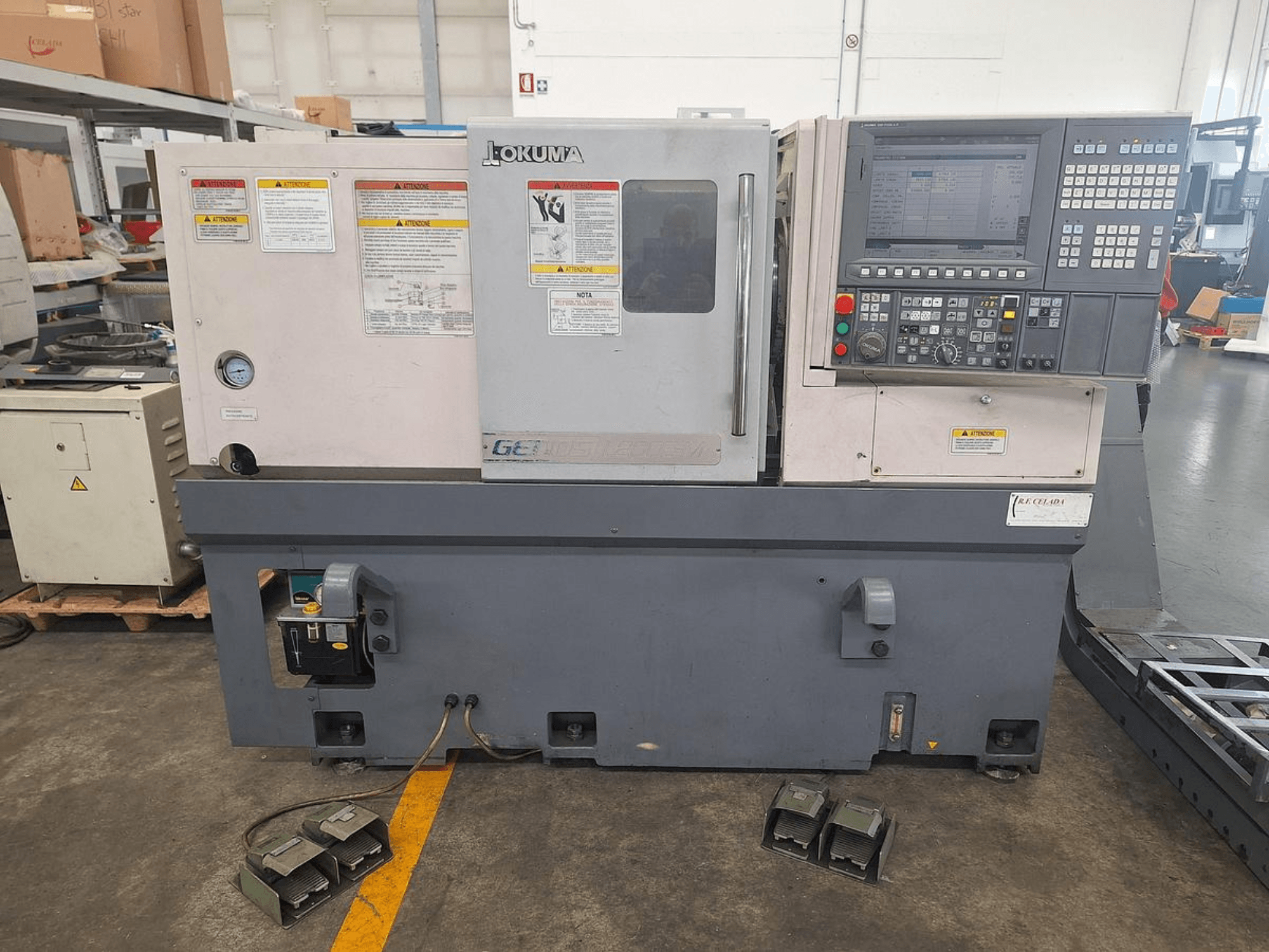 Okuma GENOS L200E-M vízszintes esztergagép oldalnézetben; vezérlőpanel, biztonsági figyelmeztetések és lábpedálok.