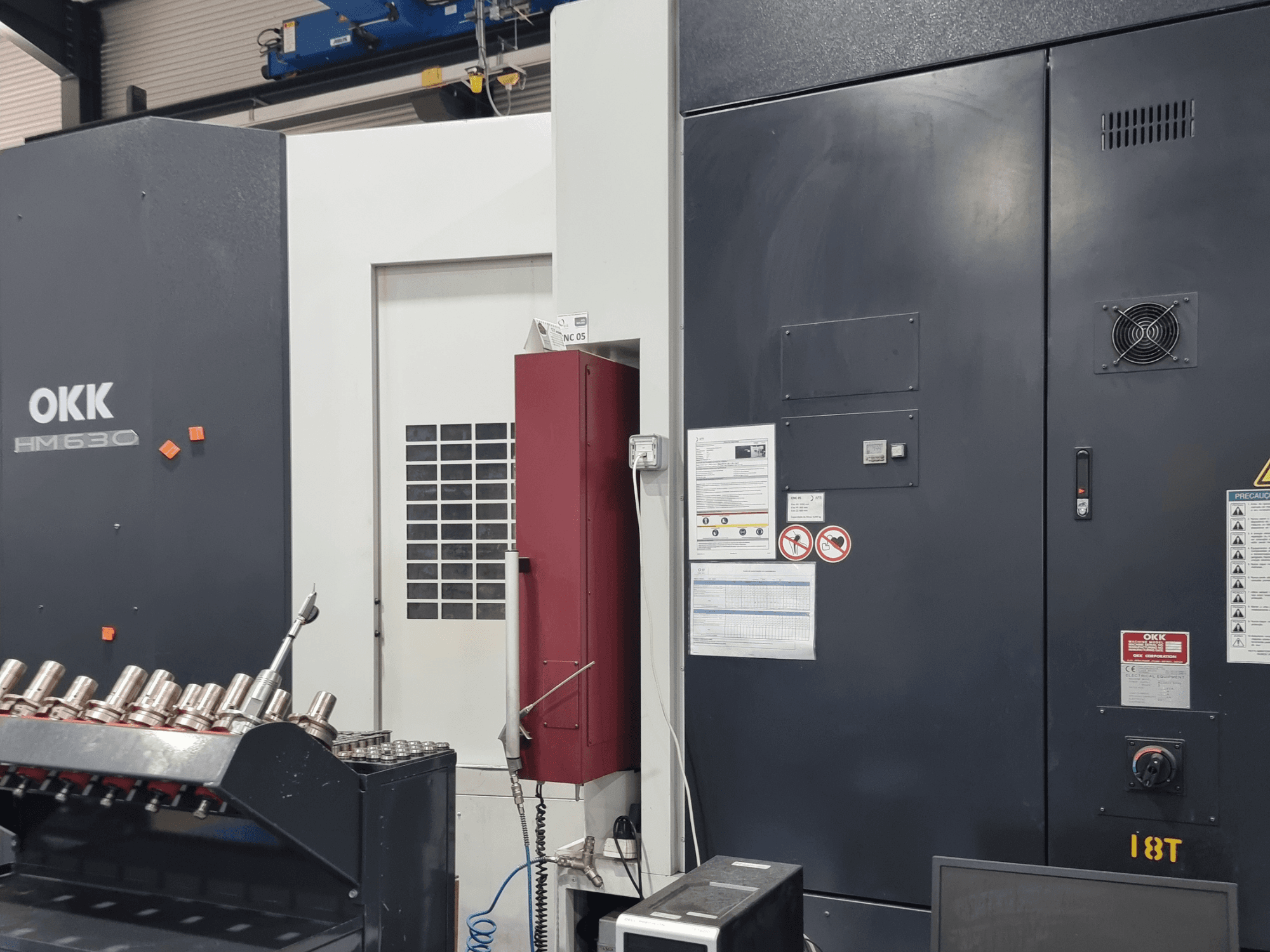 OKK HM630 vízszintes megmunkálóközpont, elölnézet, amelyen látható a vezérlőpanel, a biztonsági figyelmeztetések és az oldalán elhelyezett szerszámtároló.