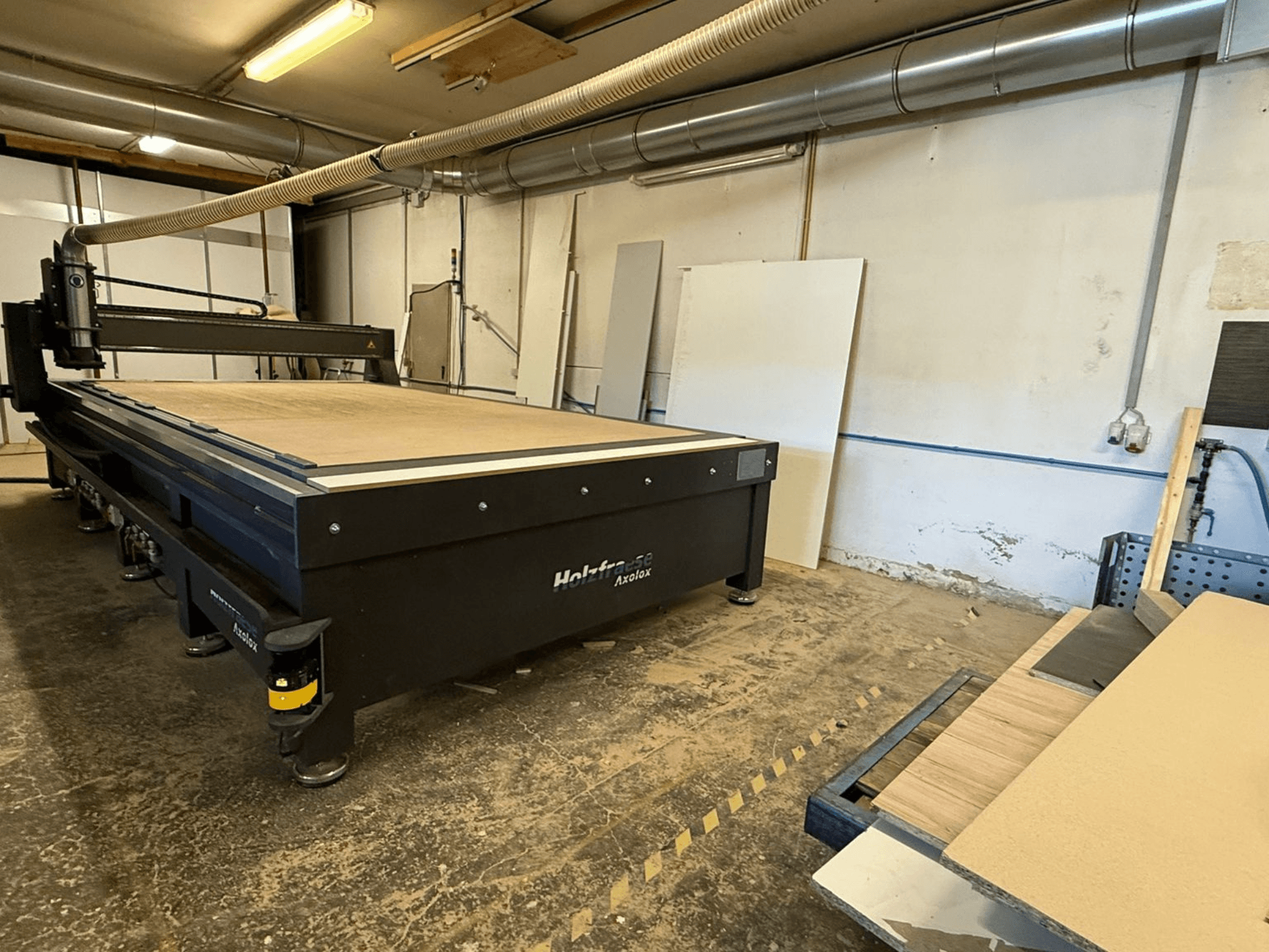 Holz-Her Axos 5 CNC-router a műhelyben, elölnézetben a munkaasztal, a porelszívó rendszer és a közeli falapok.