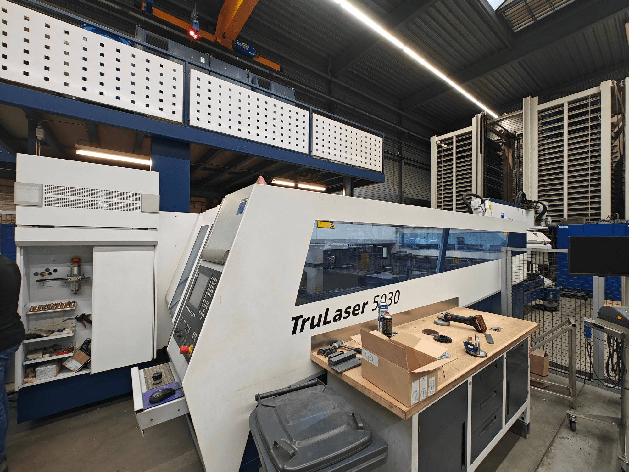 TruLaser 5030 gép egy műhelyben, elölnézetben, a vezérlőpanel látható, szerszámokkal és berendezésekkel körülvéve.