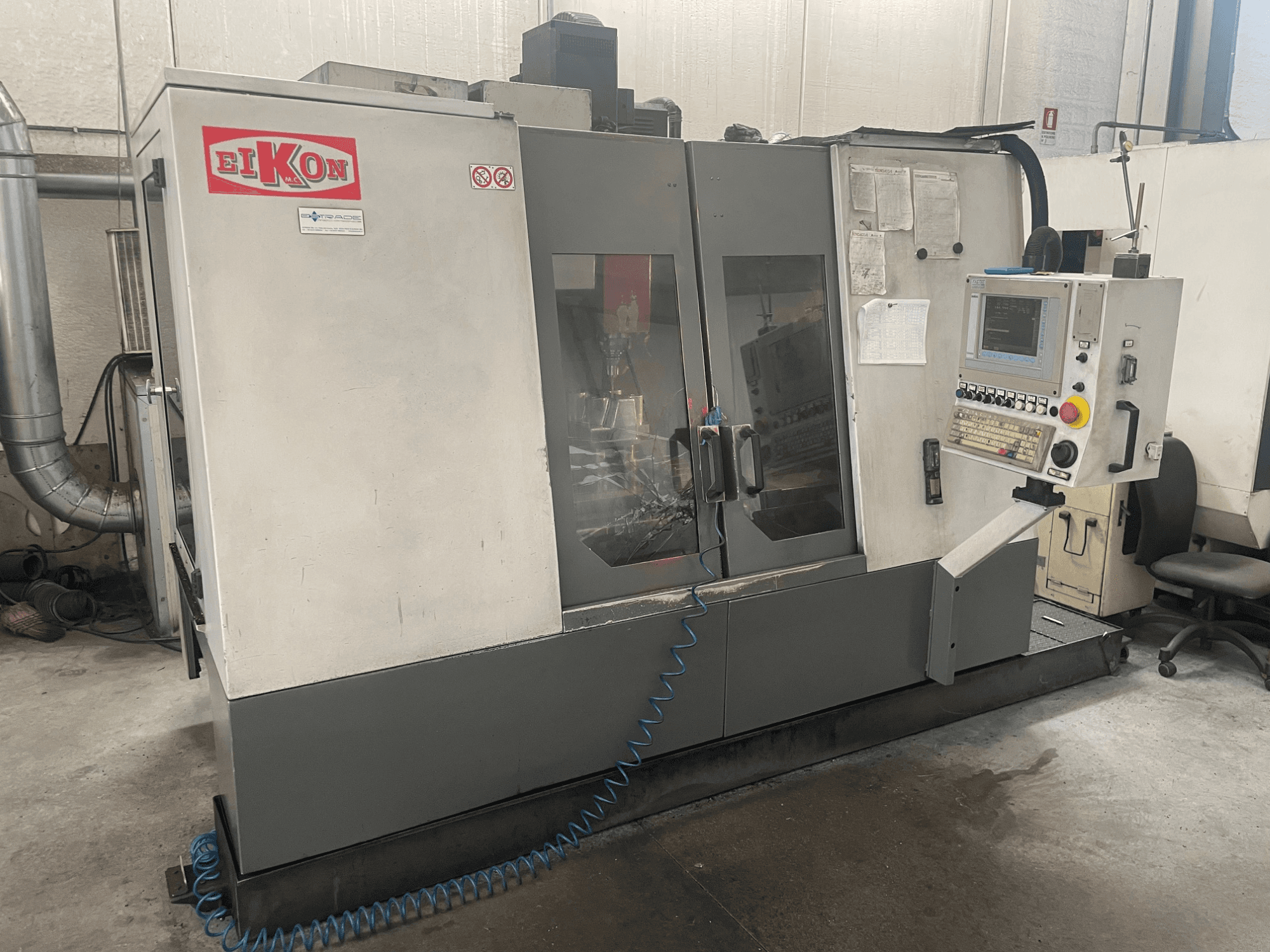 EIKON CNC-gép elölről nézve, a vezérlőpanel, a nagy üvegajtók és a kék légtömlő a padlón.