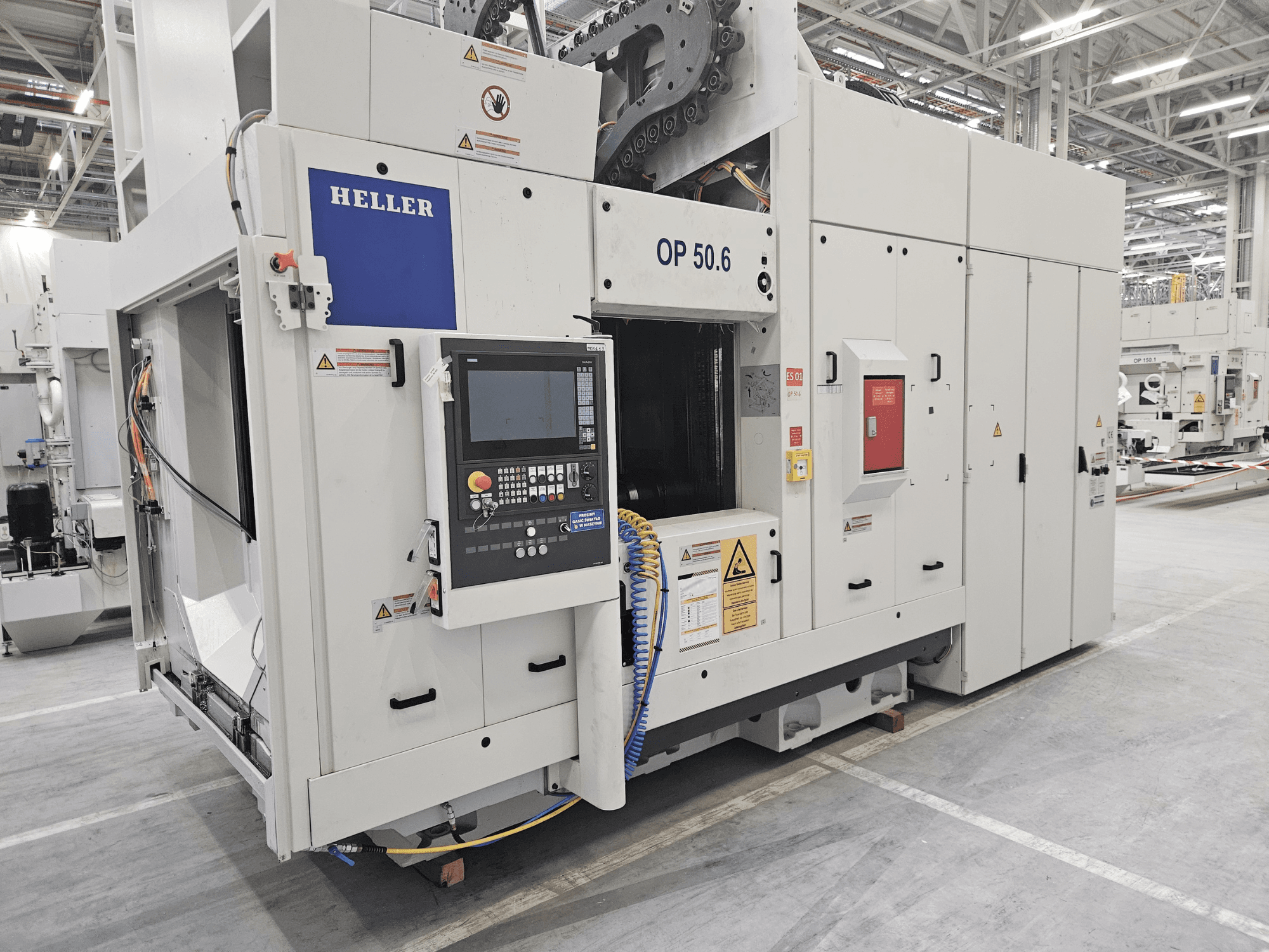 Heller OP 50.6 megmunkálóközpont, elölnézet, a vezérlőpanel, a figyelmeztető címkék és a tömlők láthatóak gyári környezetben.