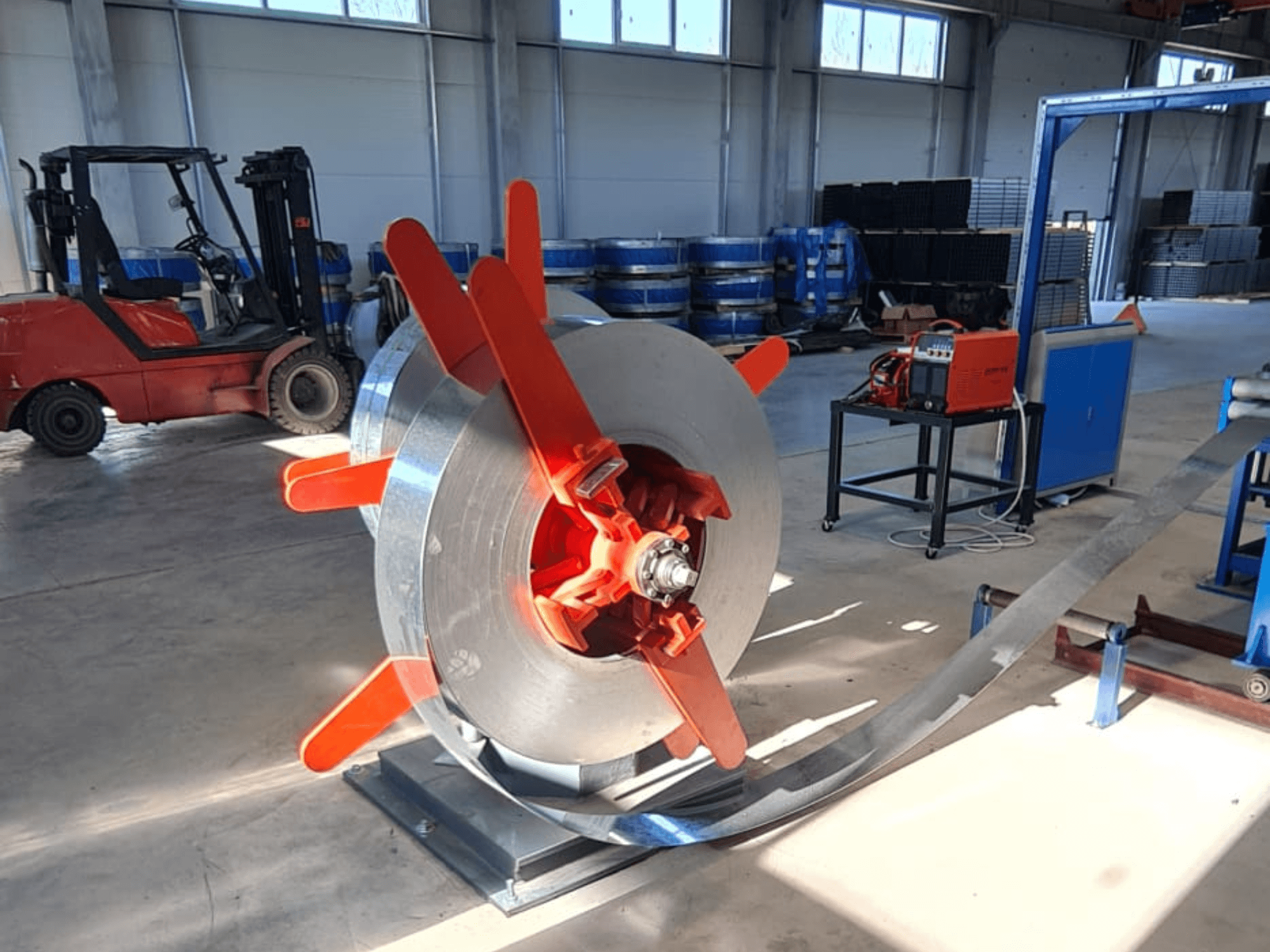 Fémtekercs piros fogantyúval állványon, szögből nézve. Ipari háttér gépekkel és targoncával.