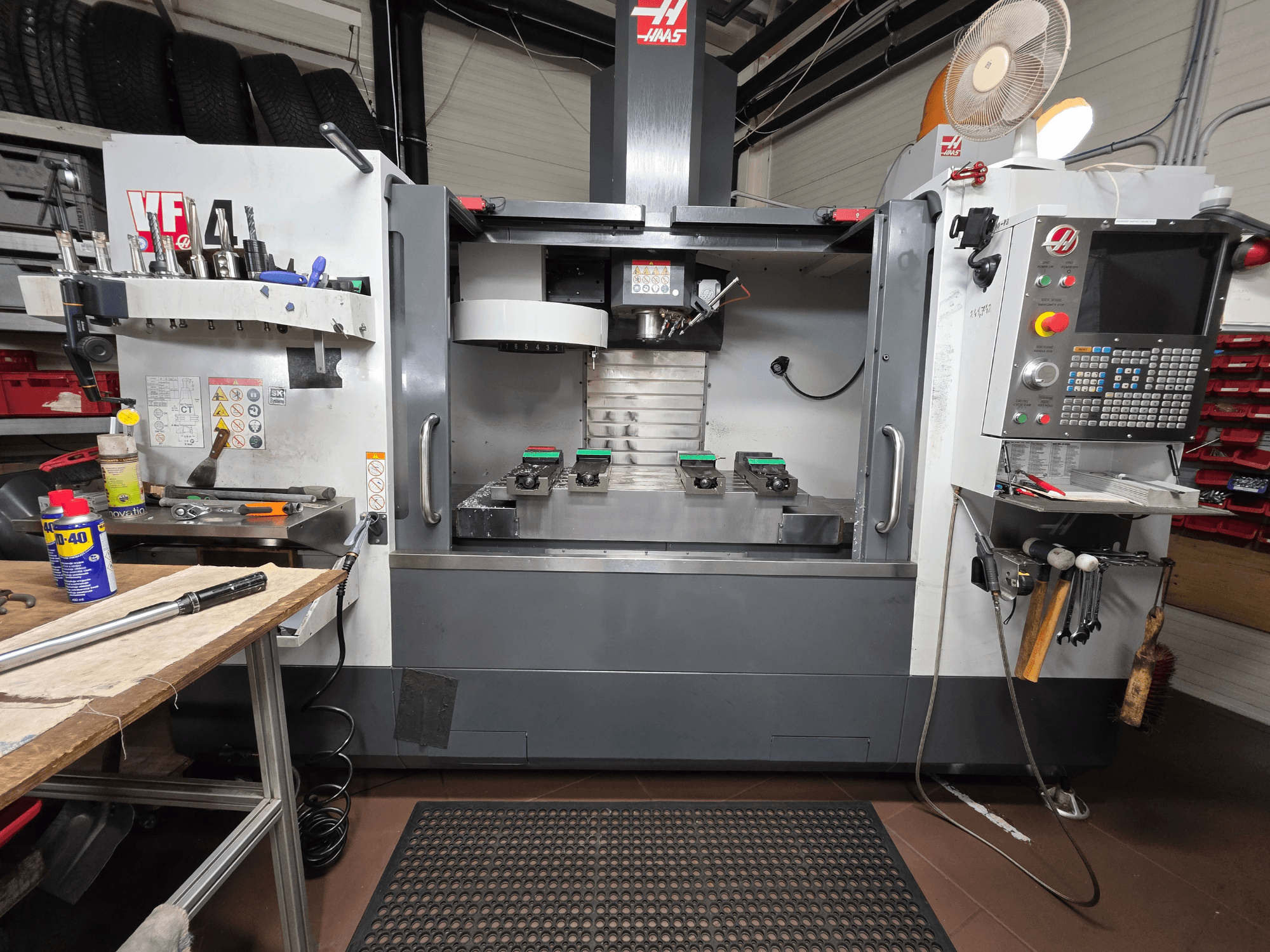 Haas VF-4 CNC-malom elölnézetben egy műhelyben, szerszámokkal, számítógépes interfésszel és két munkadarabbal az asztalon.