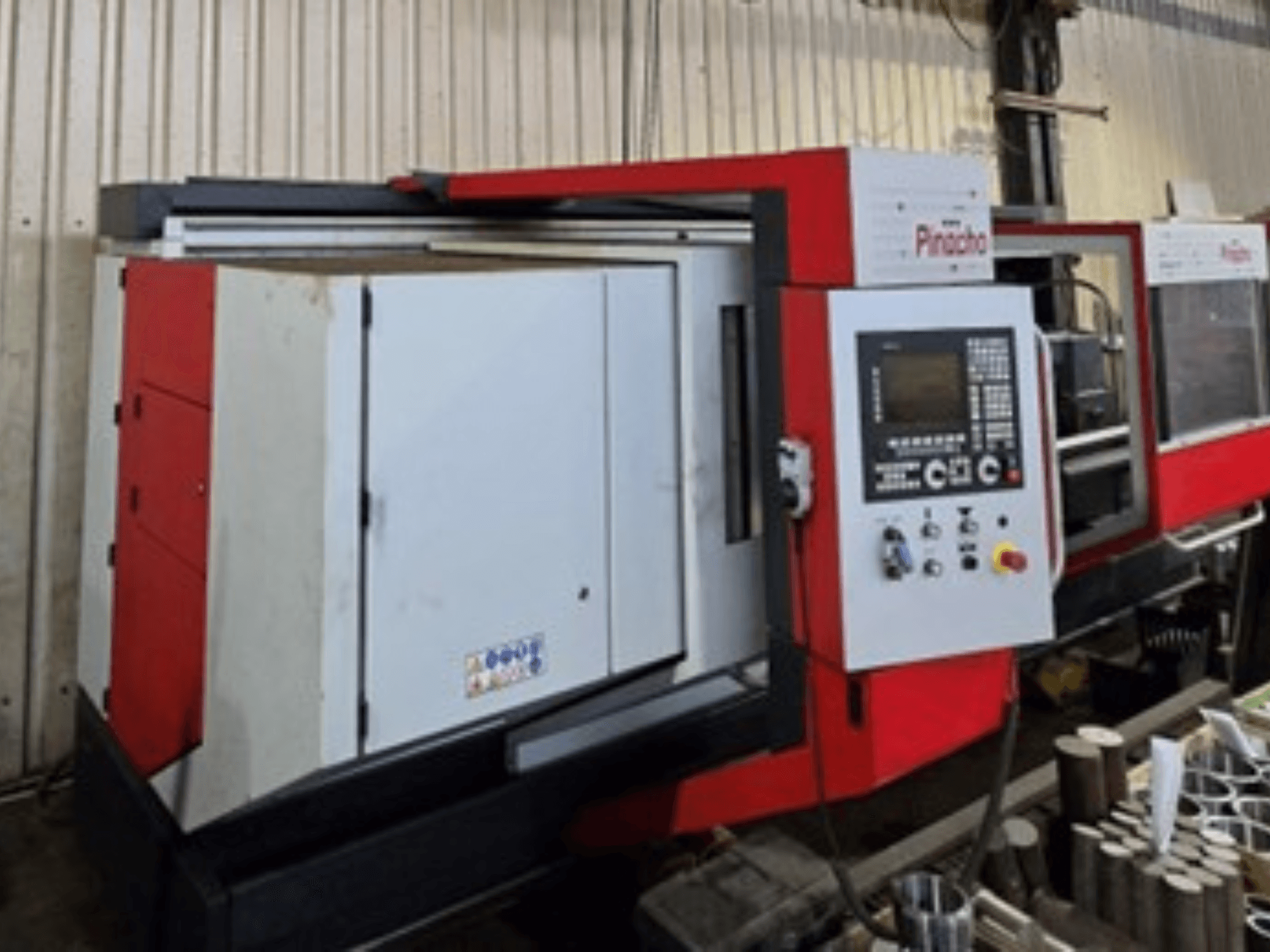 Pinacho CNC-gép piros-fehér színben, oldalnézetben, a vezérlőpanellel és a padlón lévő anyagokkal, működési beállításokat mutat.