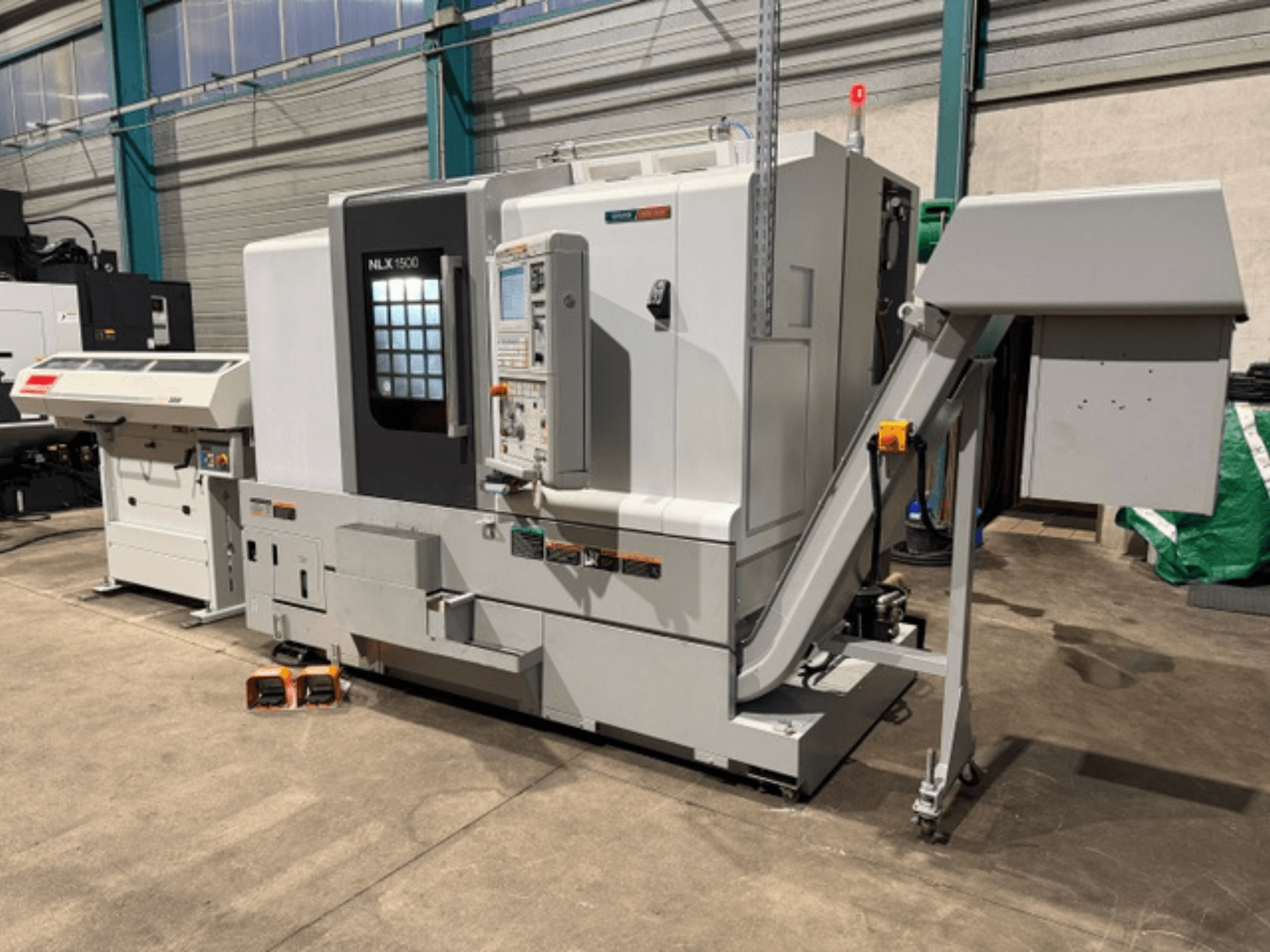 NLK 1500 CNC gép, elölnézet, vezérlőpanel és tartozéktartály az oldalon, betonpadlóra támasztva.
