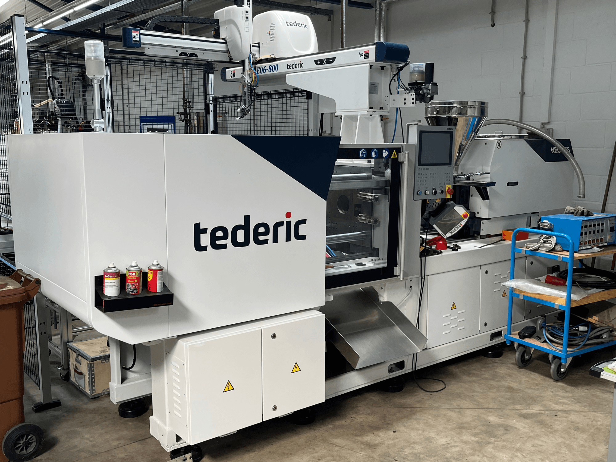 TEDERIC NEO.E55/e110h hidraulikus fröccsöntőgép egy műhelyben, oldalnézet a vezérlőpanellel és a mellette elhelyezett szerszámokkal.