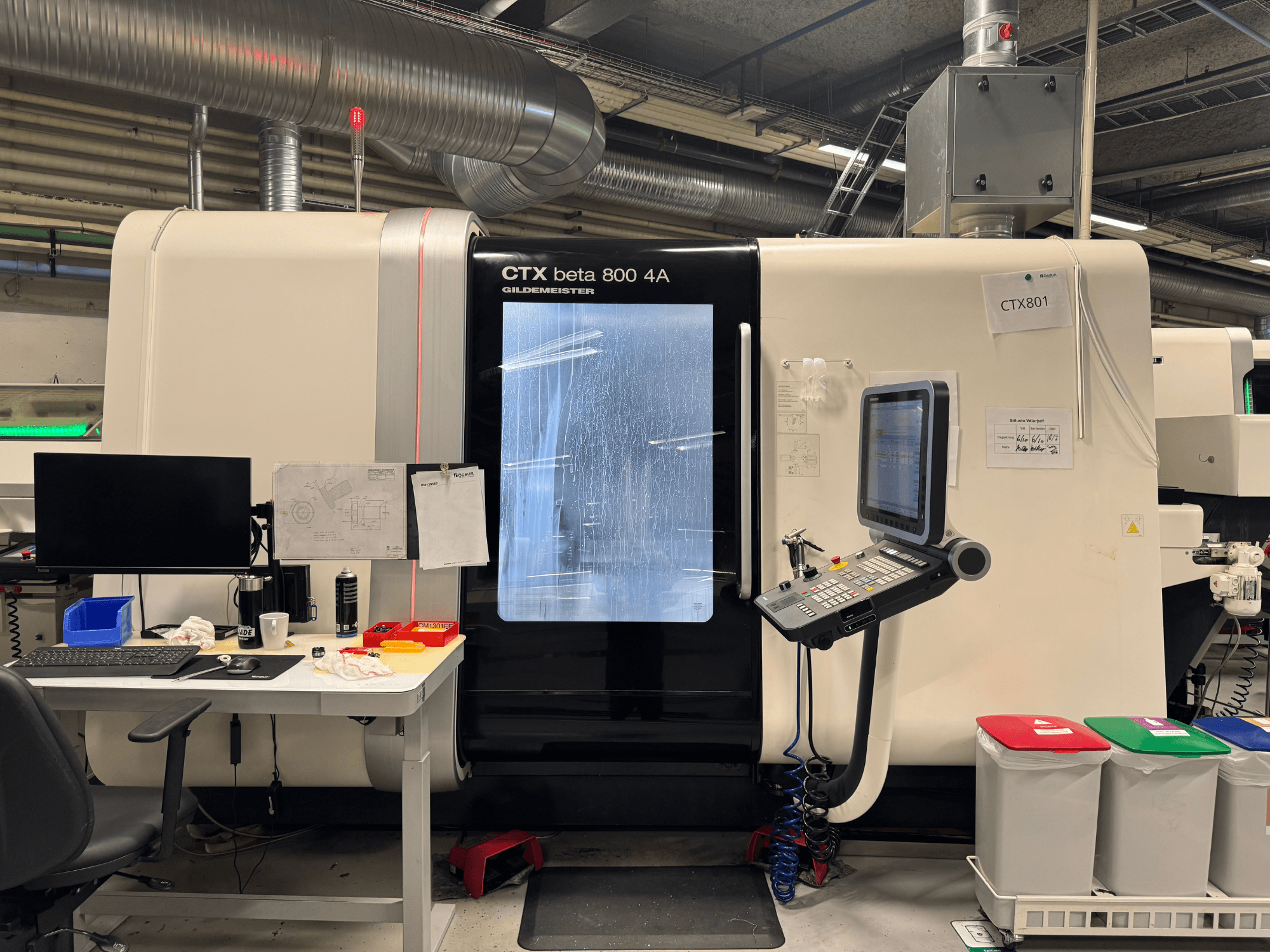 CTX beta 800 4A gép vezérlőpanellel és monitorral, oldalnézetben, egy műhelyben, irodai berendezéssel és színes szemetesekkel.