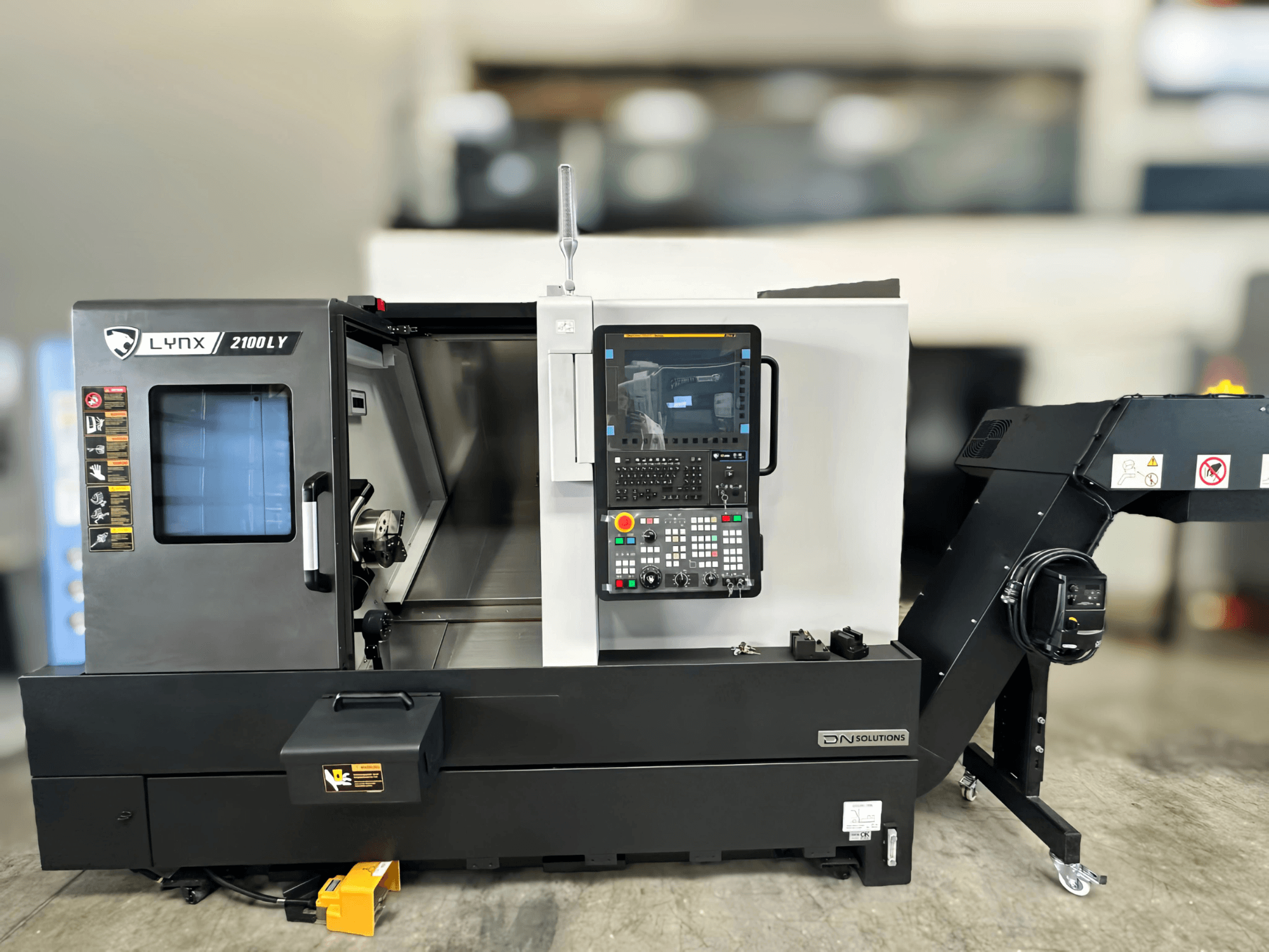 Vízszintes esztergagép DN SOLUTIONS LYNX 2100LYB, elölnézet, a kezelőpanel és a nyitott ajtó, amely a belső teret mutatja.