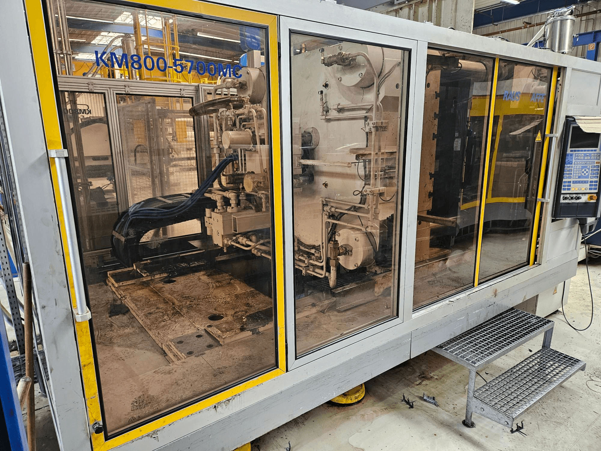 Krauss Maffei KM800-5700 MC hidraulikus fröccsöntőgép, elölnézet, amelyen az üvegpanelen keresztül láthatók az alkatrészek.