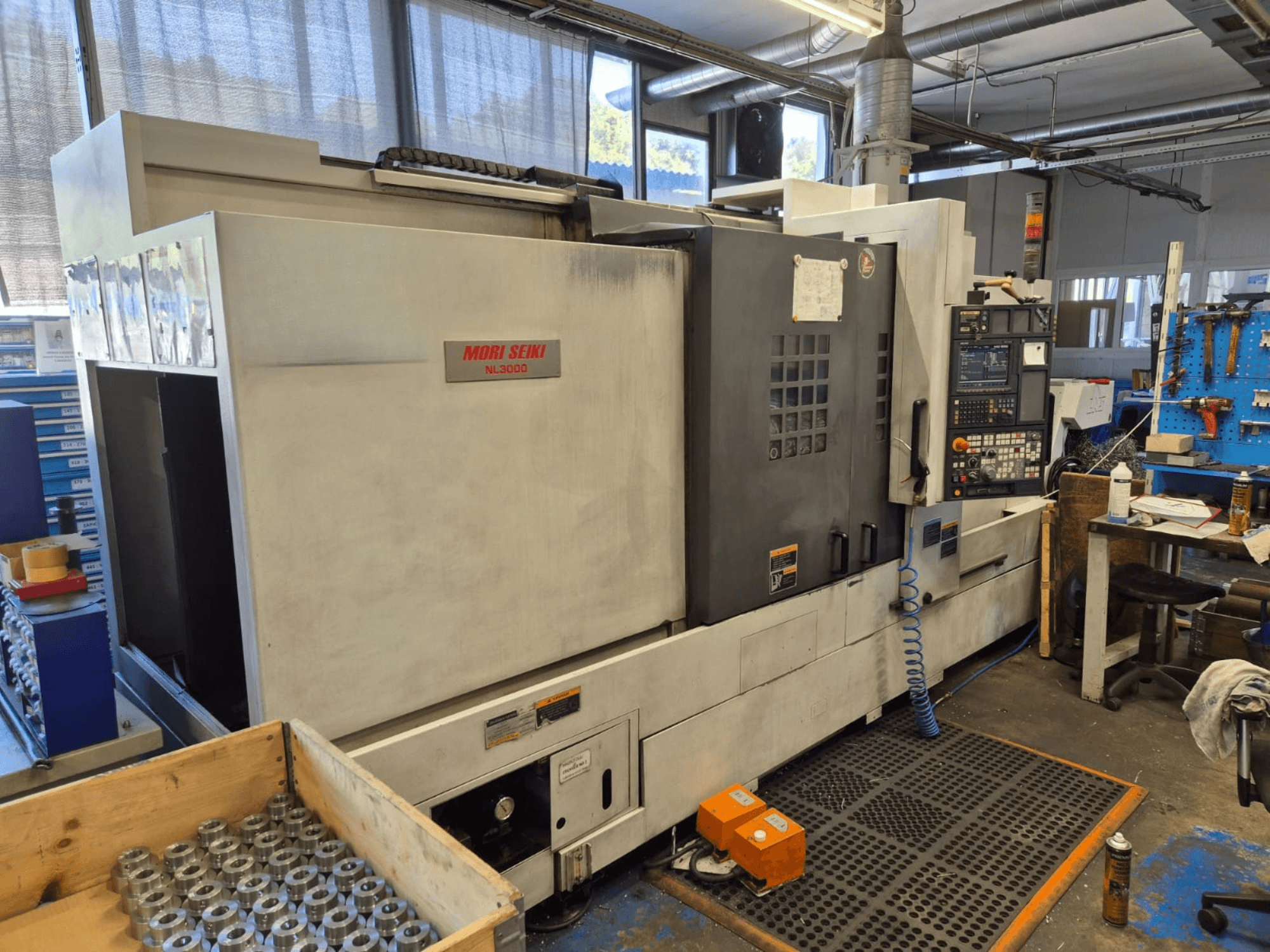 Mori Seiki NL3000 CNC esztergagép egy műhelyben, oldalnézetben a vezérlőpanel, a szerszámok és a fém alkatrészek egy fadobozban.