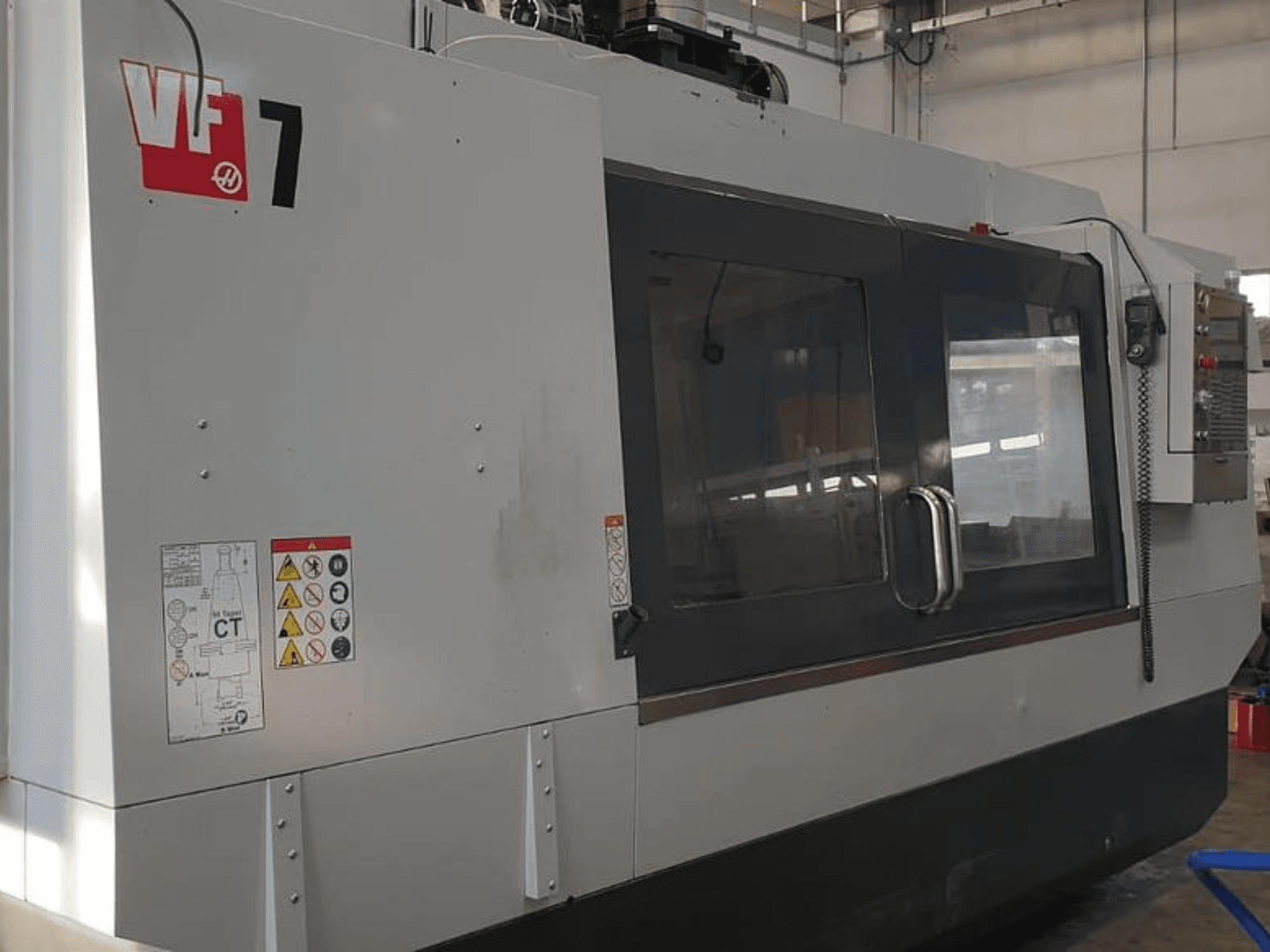 Haas VF-7 CNC-gép oldalnézetben, fémszürke kivitelben, vezérlőpanel és biztonsági címkék.