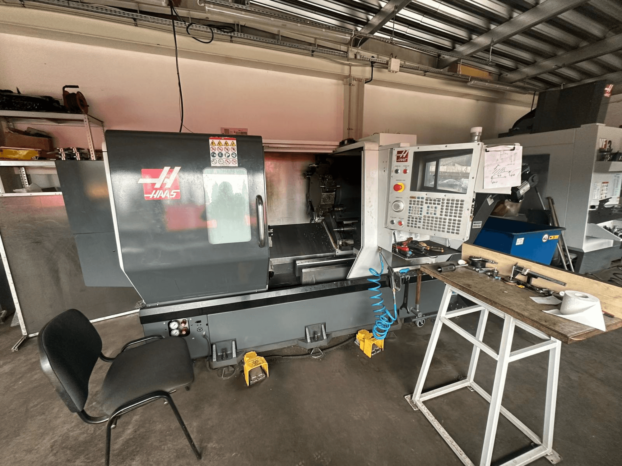 Haas CNC eszterga (ST-10 modell) egy műhelyben, oldalnézetben a kezelőszervek, a szerszámok a munkapadon és egy szék a közelben.