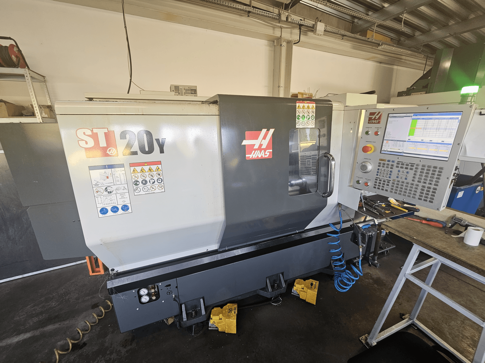 Haas ST-20Y CNC eszterga elölnézetben, vezérlőpanellel; biztonsági címkékkel és légtömlővel az oldalán.